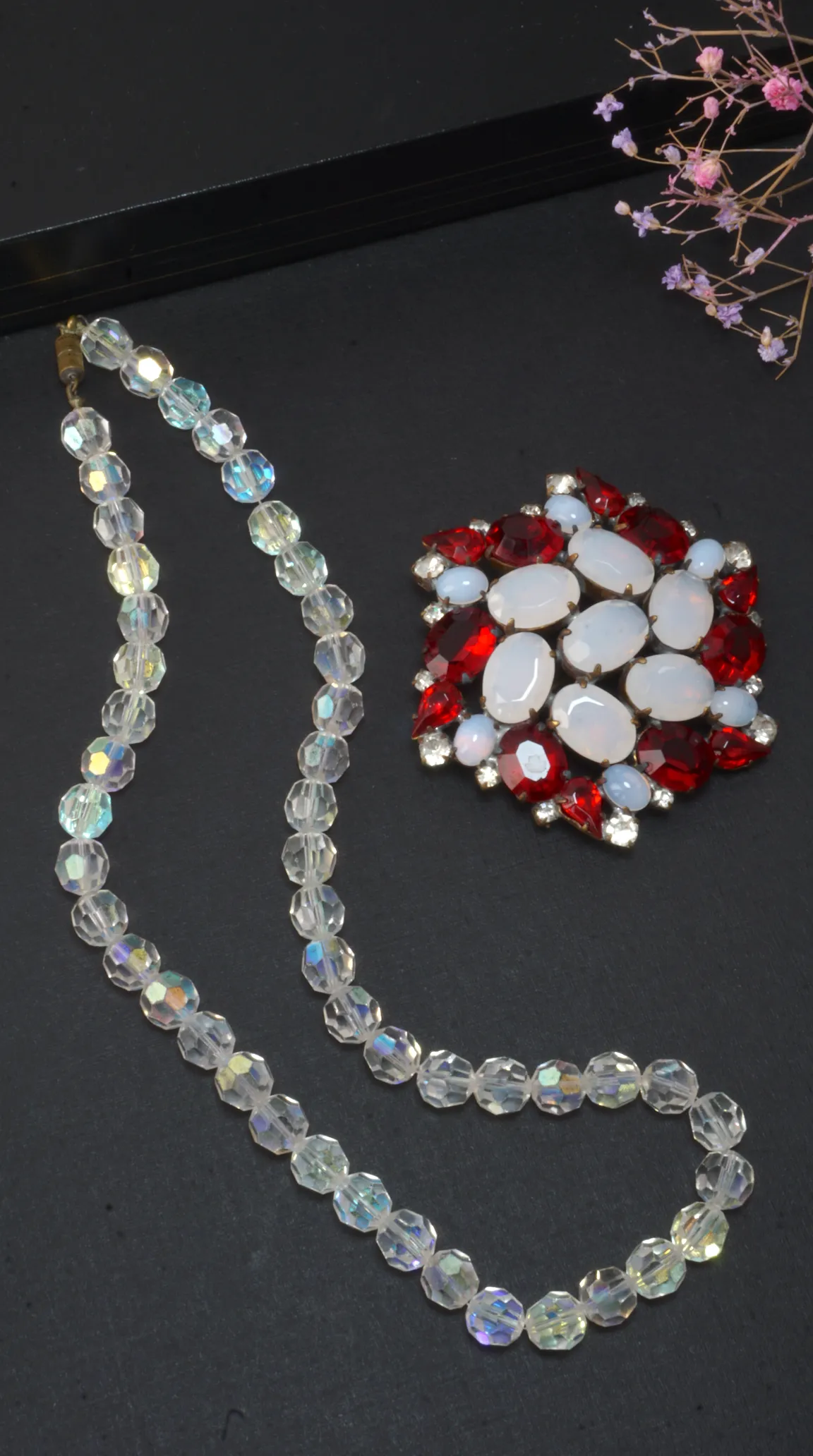 Vintage aurora borealis faceted crystal bead necklace and a red and white cabochon rhinestone brooch on a dark background.