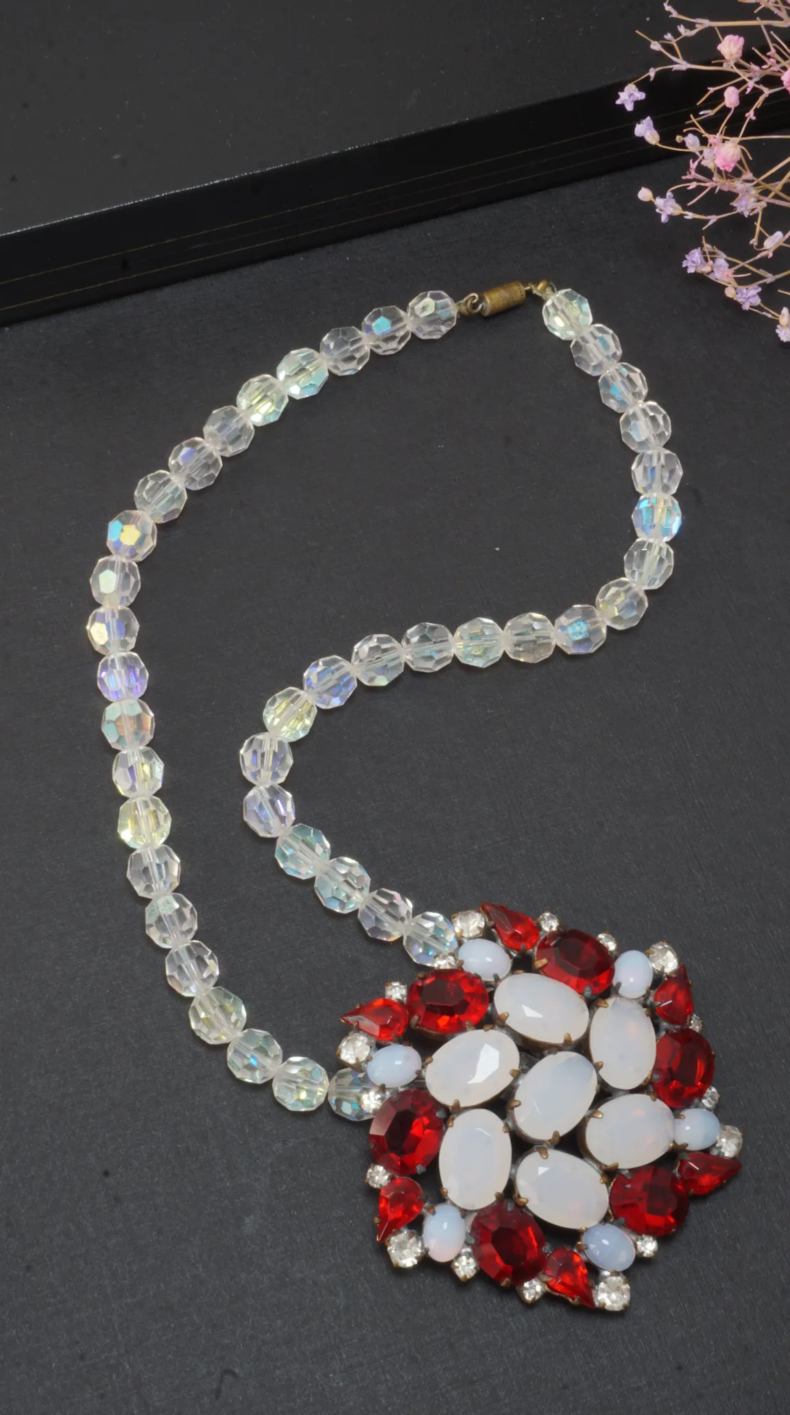 Vintage aurora borealis crystal bead necklace with ornate red rhinestone and white opal cabochon pendant on a dark background.