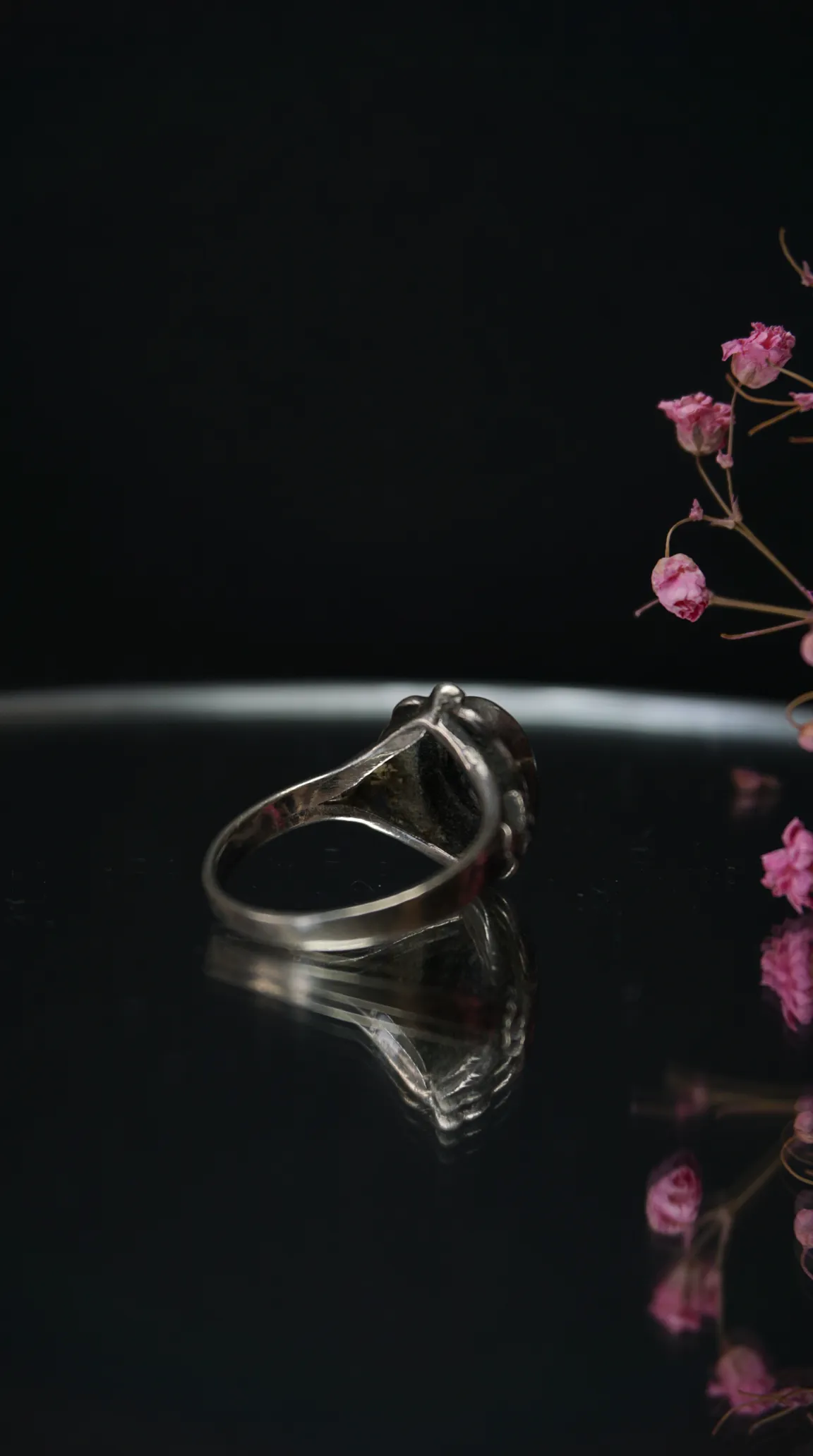Vintage silver ring with dark gemstone, side view on black reflective surface, with dried pink flowers.