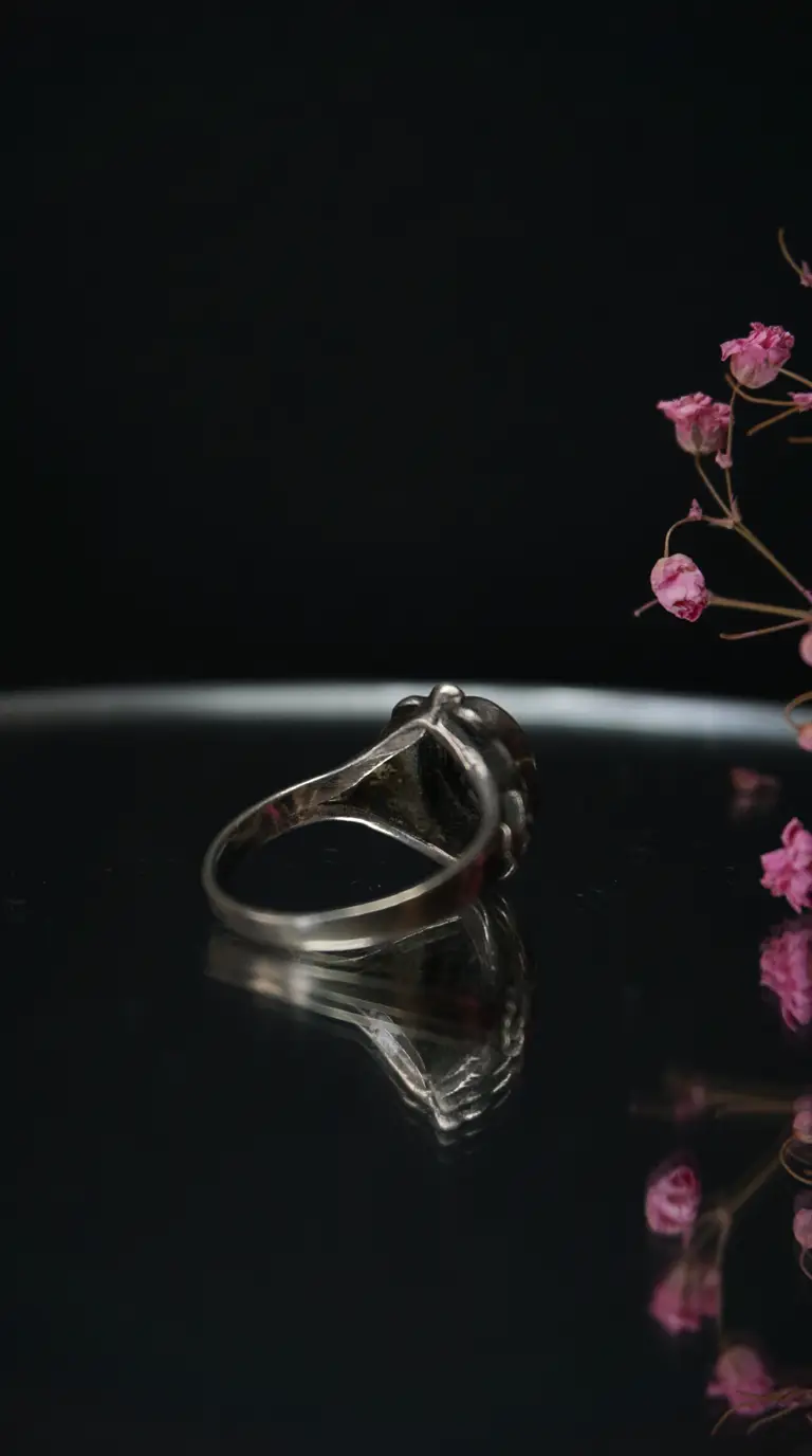 Vintage silver ring with dark gemstone, side view on black reflective surface, with dried pink flowers.