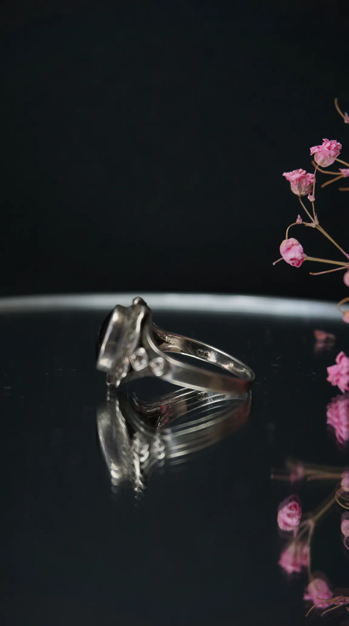 Vintage sterling silver ring with dark oval gemstone on reflective black surface, accented by pink dried flowers.