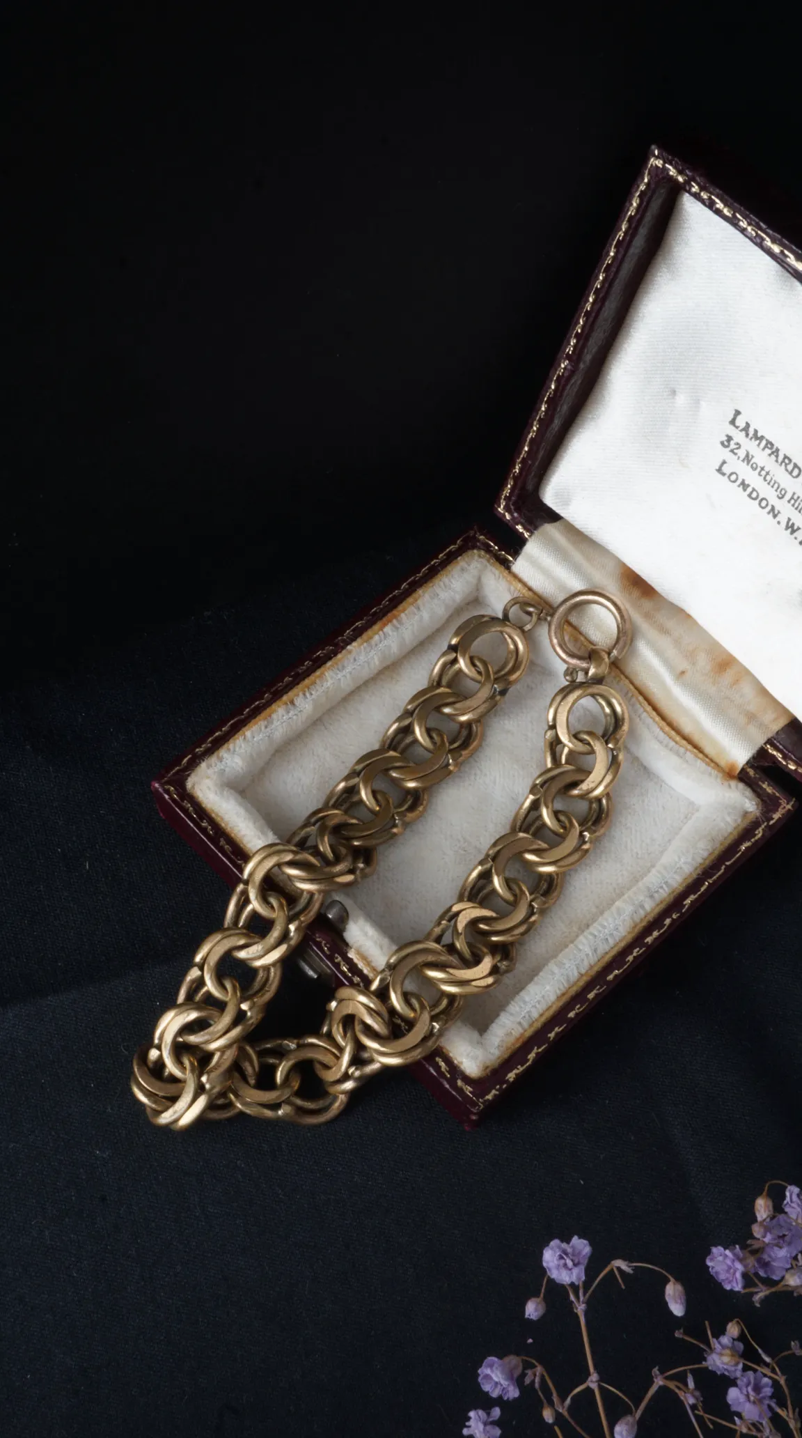 Vintage gold chain bracelet displayed in open antique Lampard London jewelry box on dark fabric with dried purple flowers.