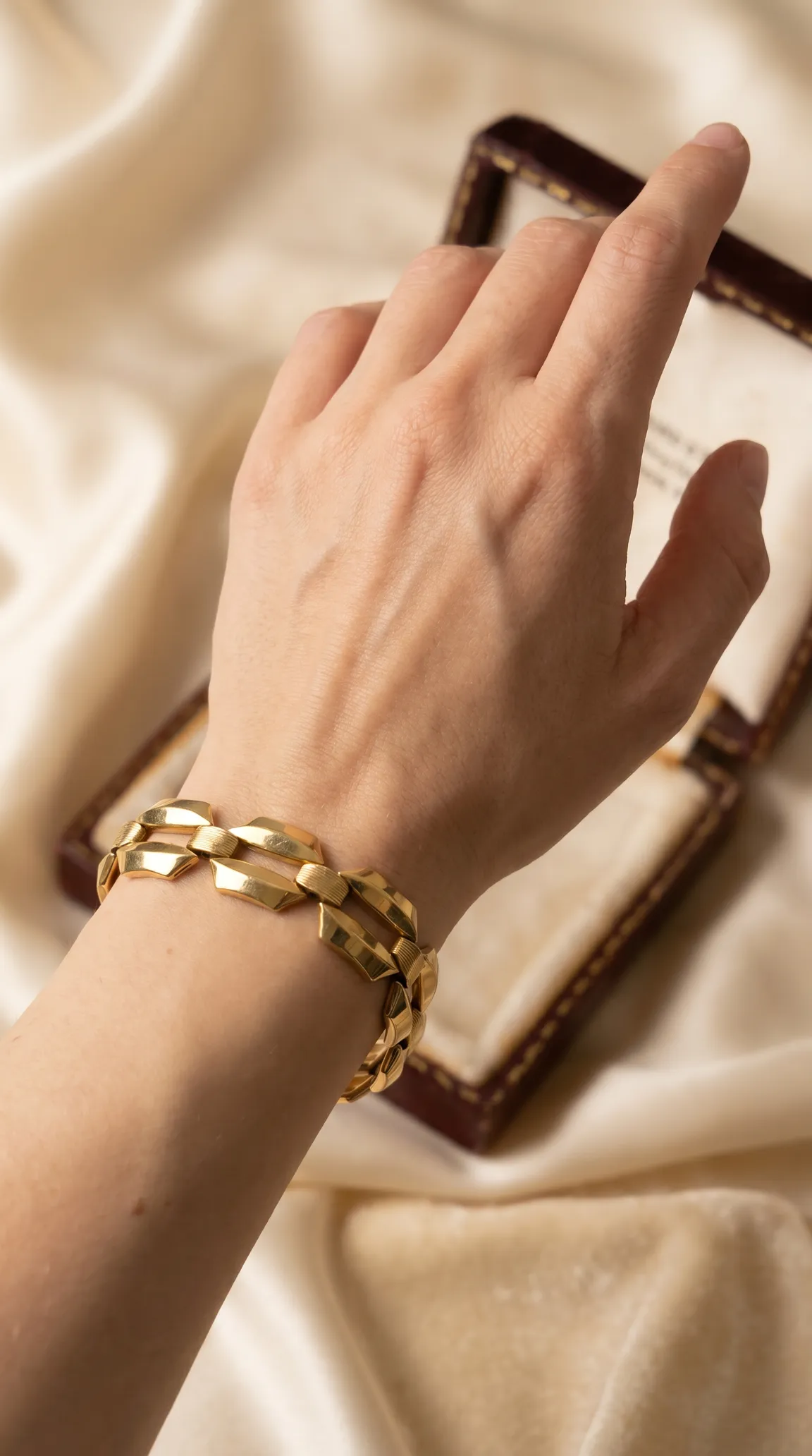 Vintage gold geometric link bracelet worn on wrist, with open jewelry box and cream silk background.