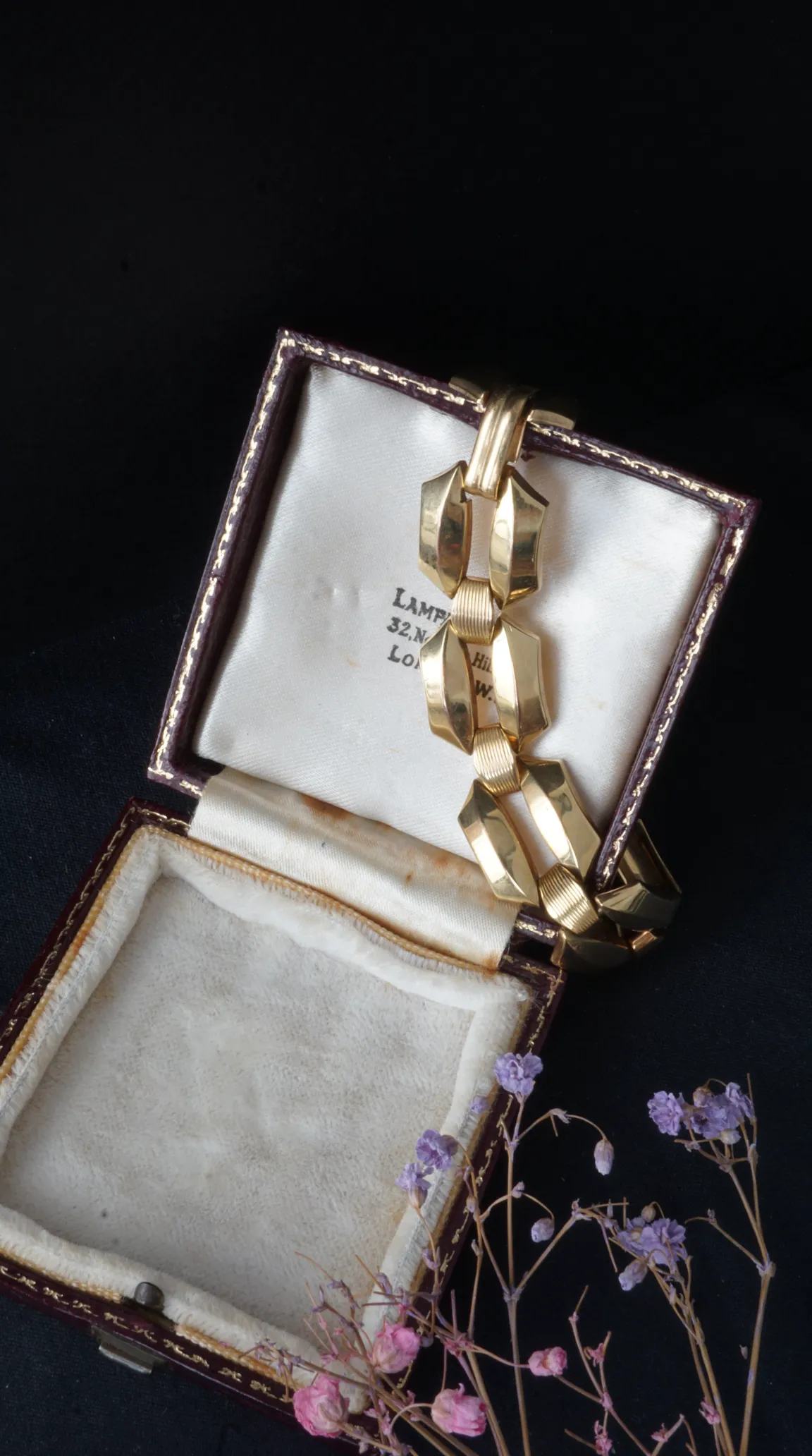 Vintage gold geometric link bracelet displayed in an open antique jewelry box with London jeweler details, accompanied by dried purple and pink flowers.