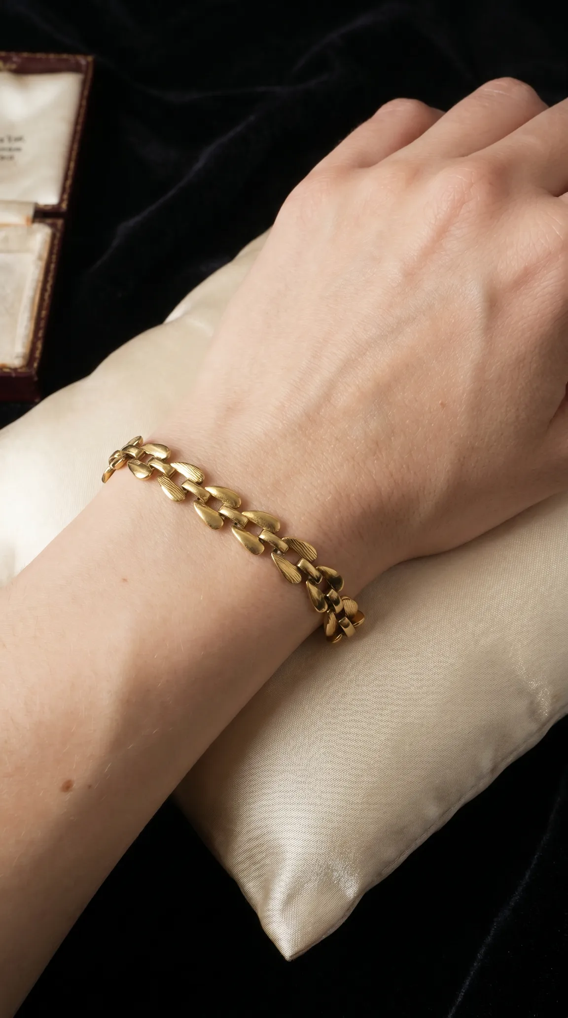 Vintage gold textured elongated link bracelet on a woman's wrist, resting on a cream satin pillow with an antique jewelry box in the background.