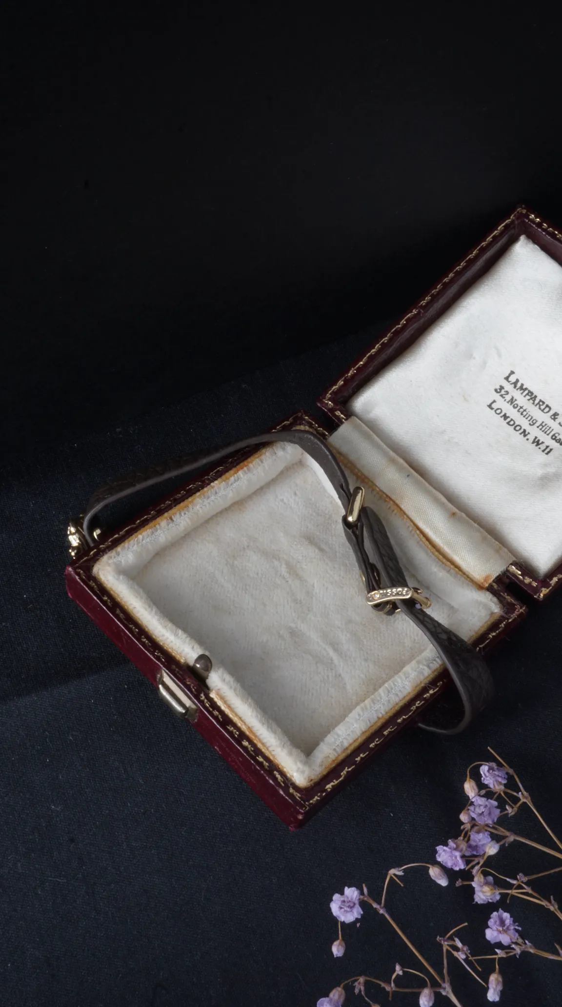 Vintage Tissot watch strap with gold and diamond buckle resting in an open antique burgundy jewelry box with cream lining and Lampard & Son, London jeweler's mark.