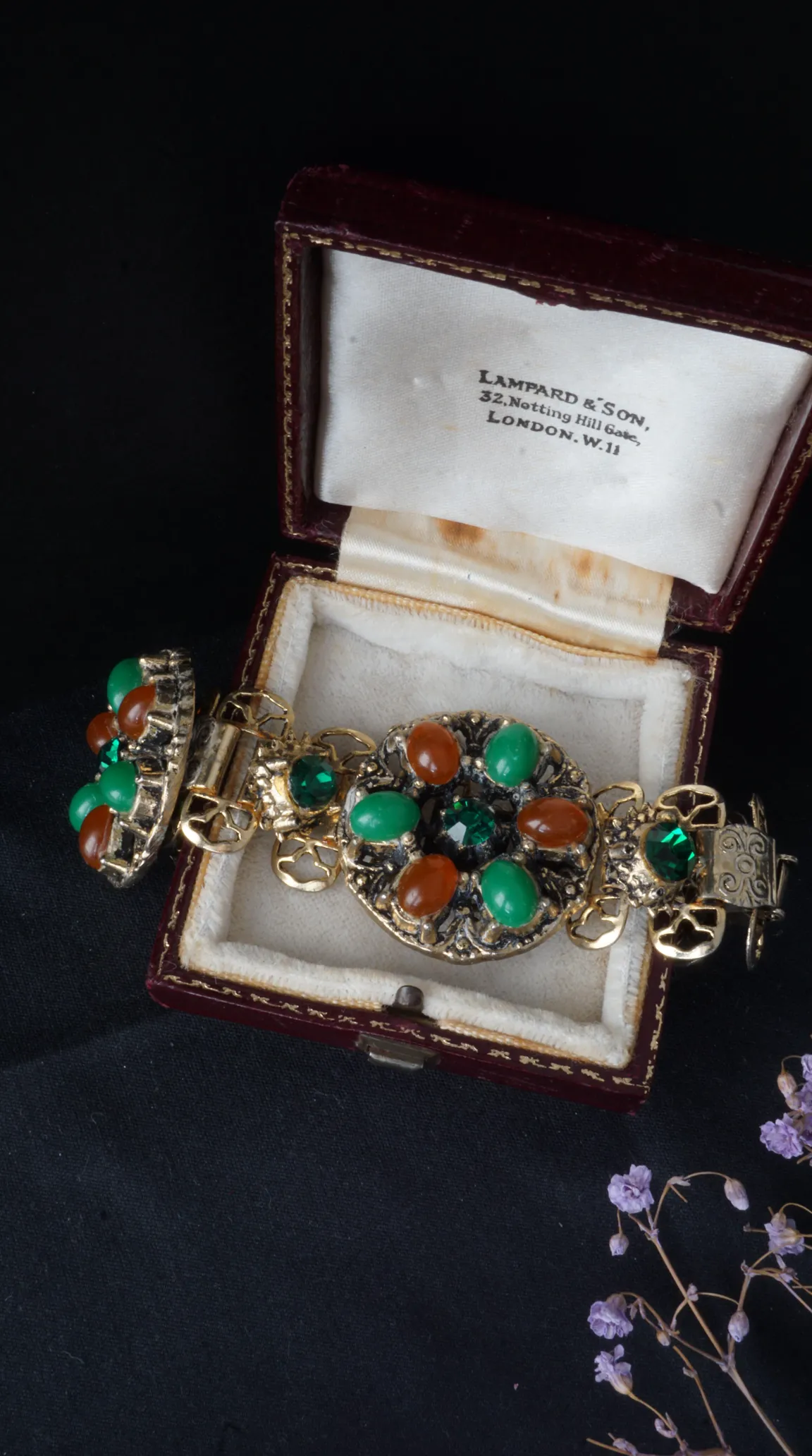 Vintage gold tone ornate bracelet and matching clip-on earrings with emerald green and amber cabochon gemstones, displayed in an open antique burgundy velvet jewelry box from Lampard & Son, London.