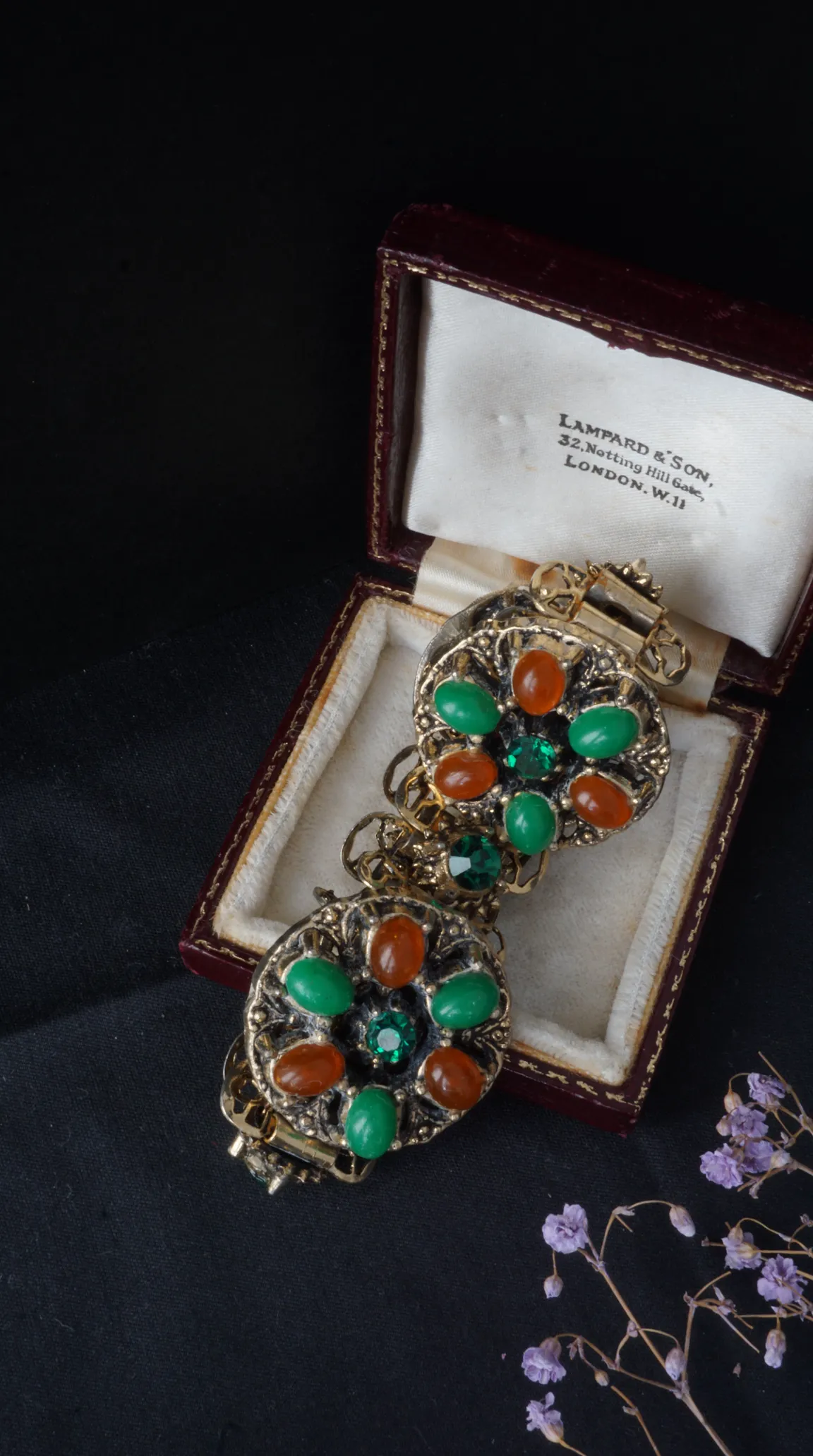 Vintage gold-tone bracelet with ornate floral design, green and orange cabochon stones, and sparkling green faceted gems, displayed in an antique red jewelry box.