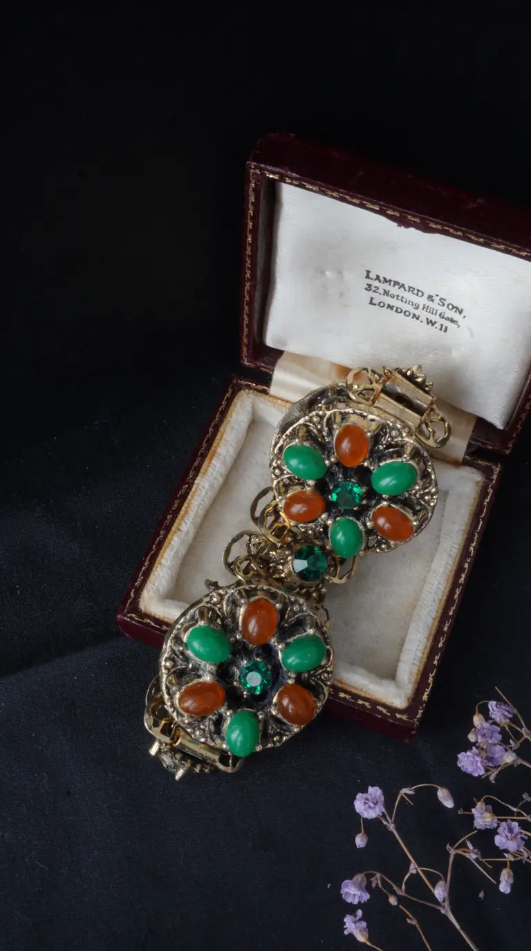 Vintage gold-tone bracelet with ornate floral design, green and orange cabochon stones, and sparkling green faceted gems, displayed in an antique red jewelry box.