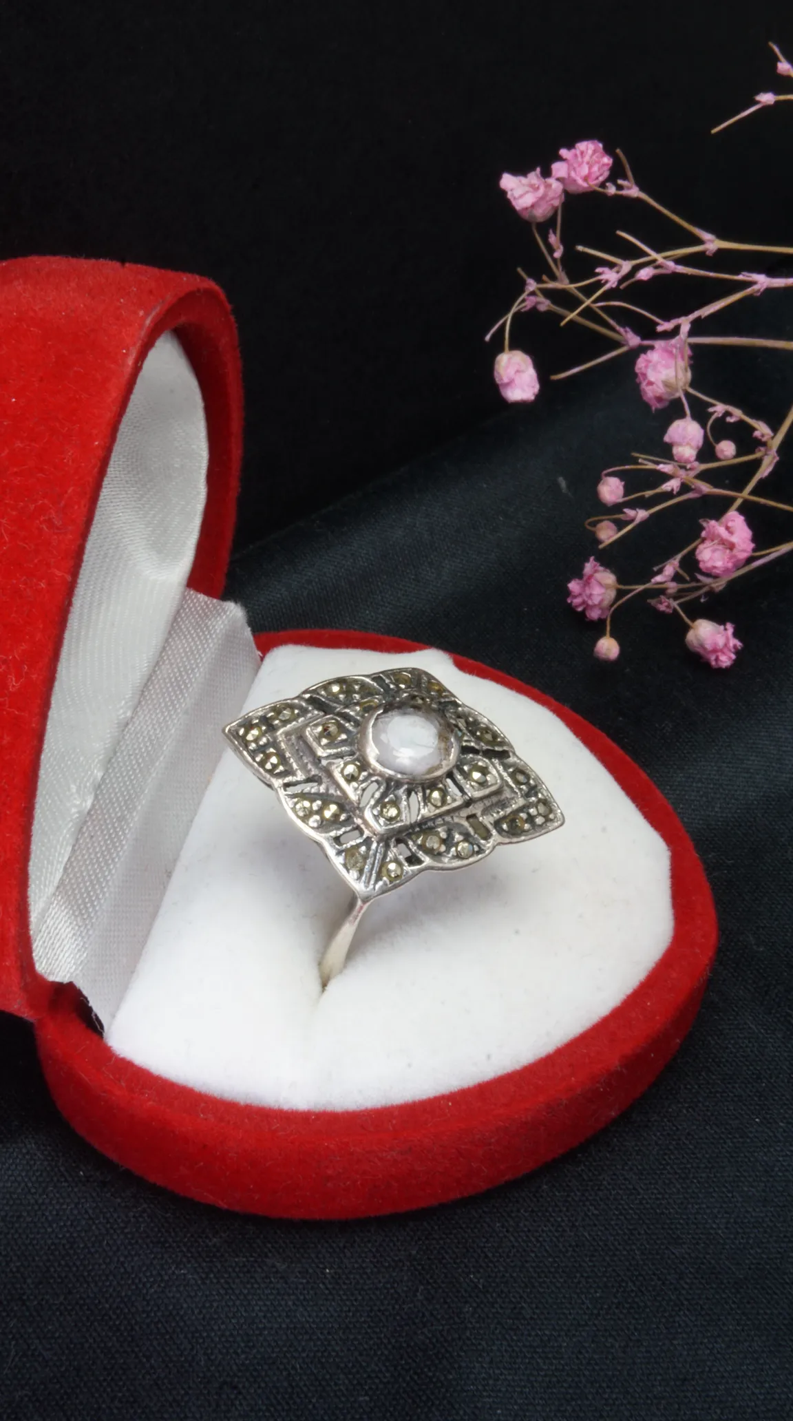 Vintage sterling silver marcasite ring with central pearl in red velvet jewelry box on black background with dried pink flowers.