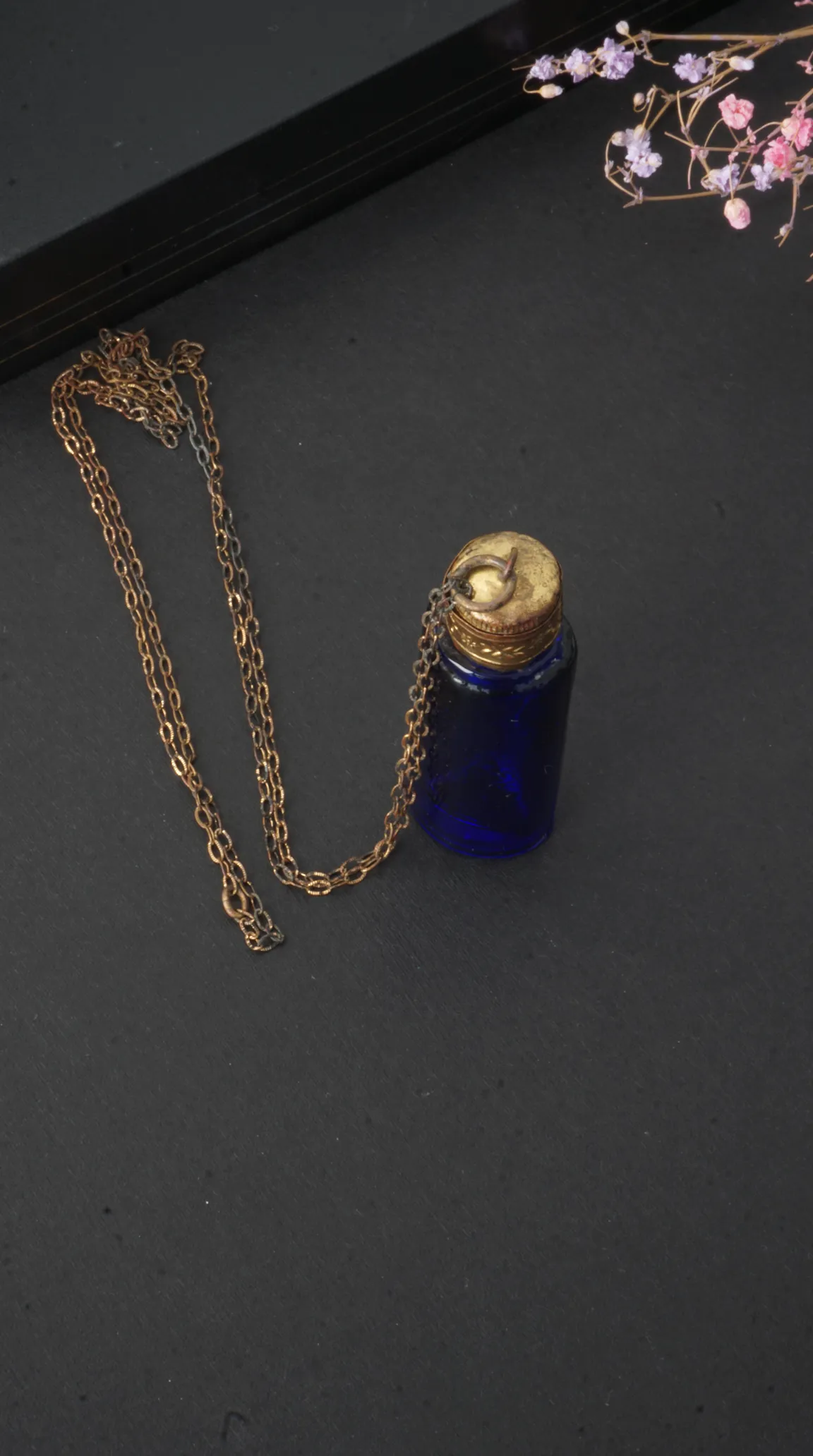 Vintage blue glass perfume bottle pendant with aged brass cap and chain on a black background with dried flowers.