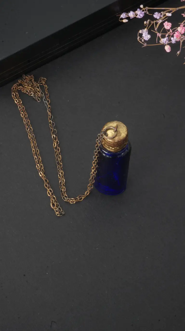 Vintage blue glass perfume bottle pendant with aged brass cap and chain on a black background with dried flowers.
