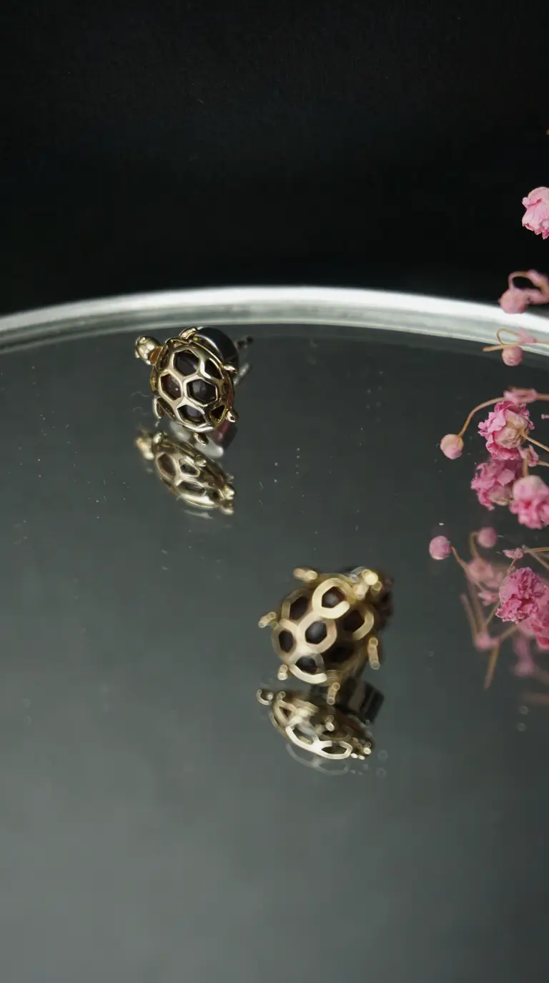 Vintage gold and black enamel turtle stud earrings reflecting on a silver surface, with dried pink flowers.