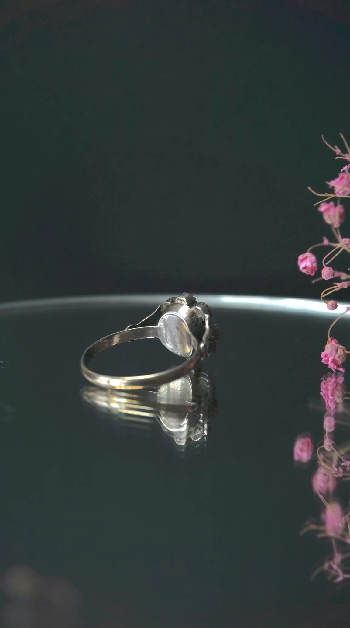 Vintage silver ring with black textured setting on reflective surface, accented by dried pink flowers.