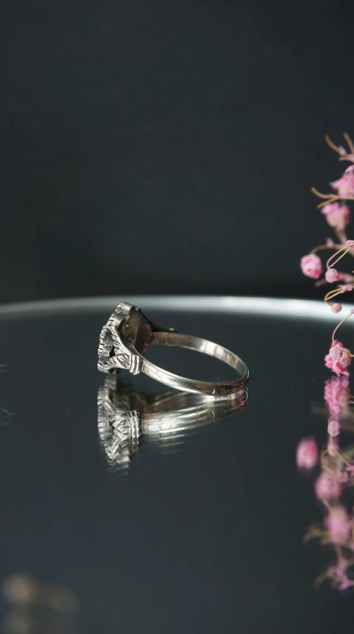 Vintage sterling silver ring with ornate details, side view on reflective surface with delicate pink flowers.