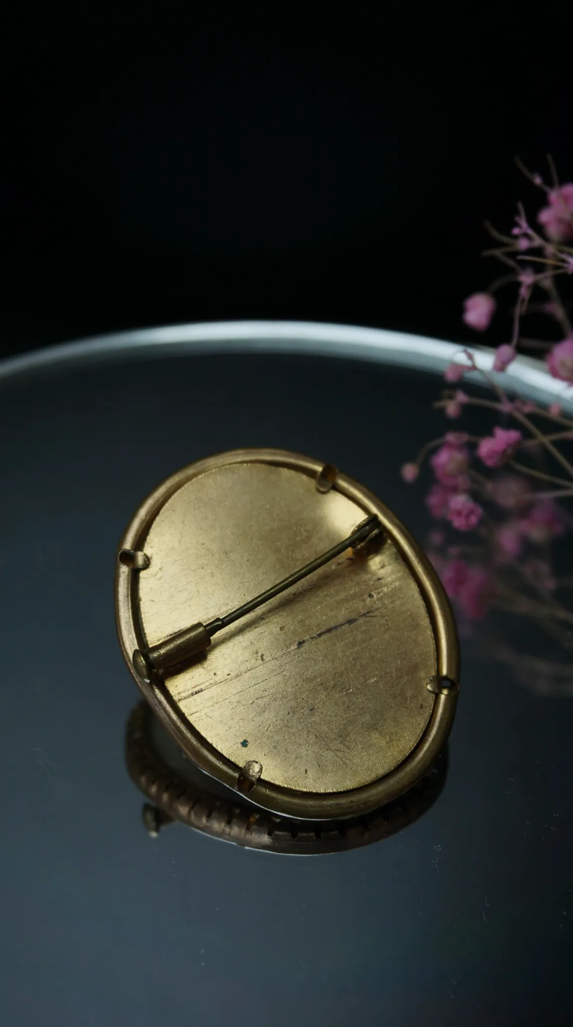 Vintage brass oval brooch back with pin clasp mechanism on reflective surface.