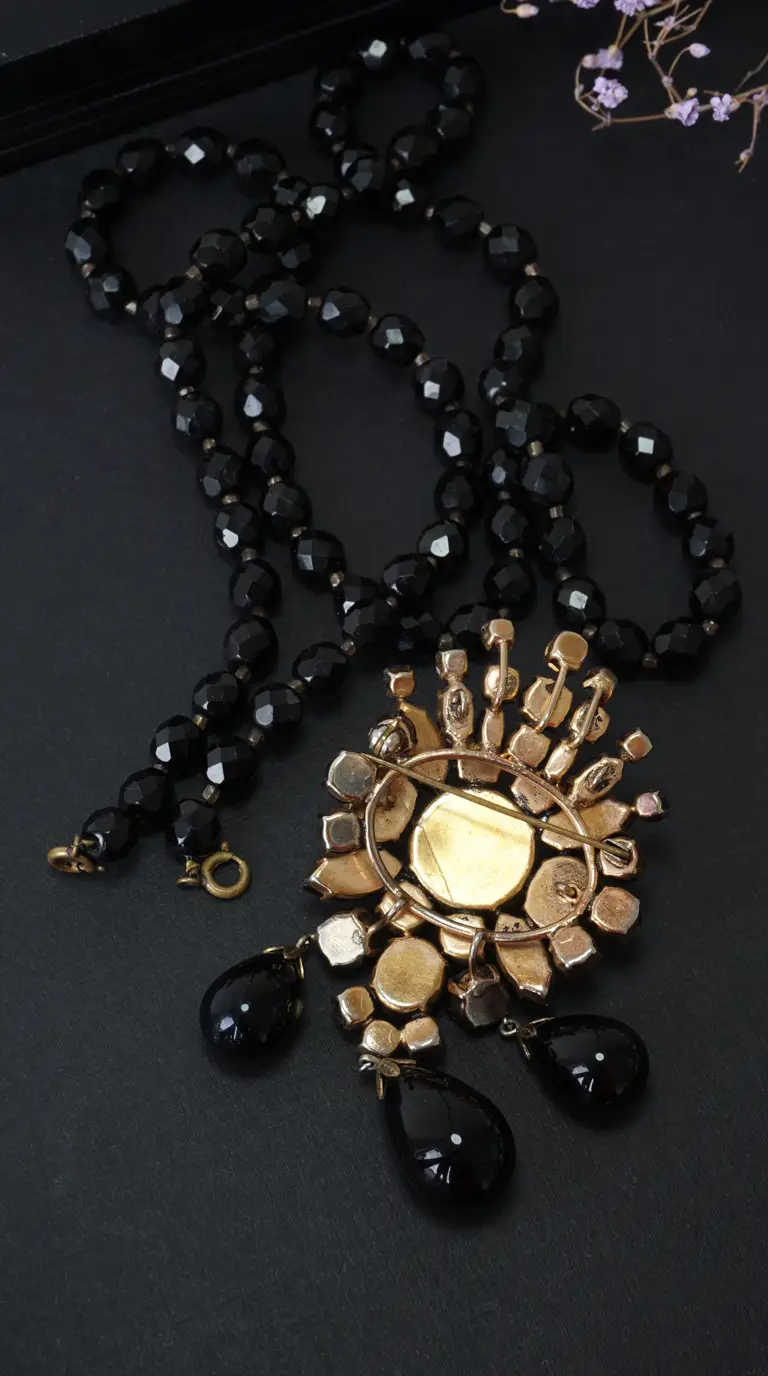 Vintage black faceted glass beaded necklace with ornate gold-tone brooch showing pin mechanism and black tear-drop dangles on dark background.