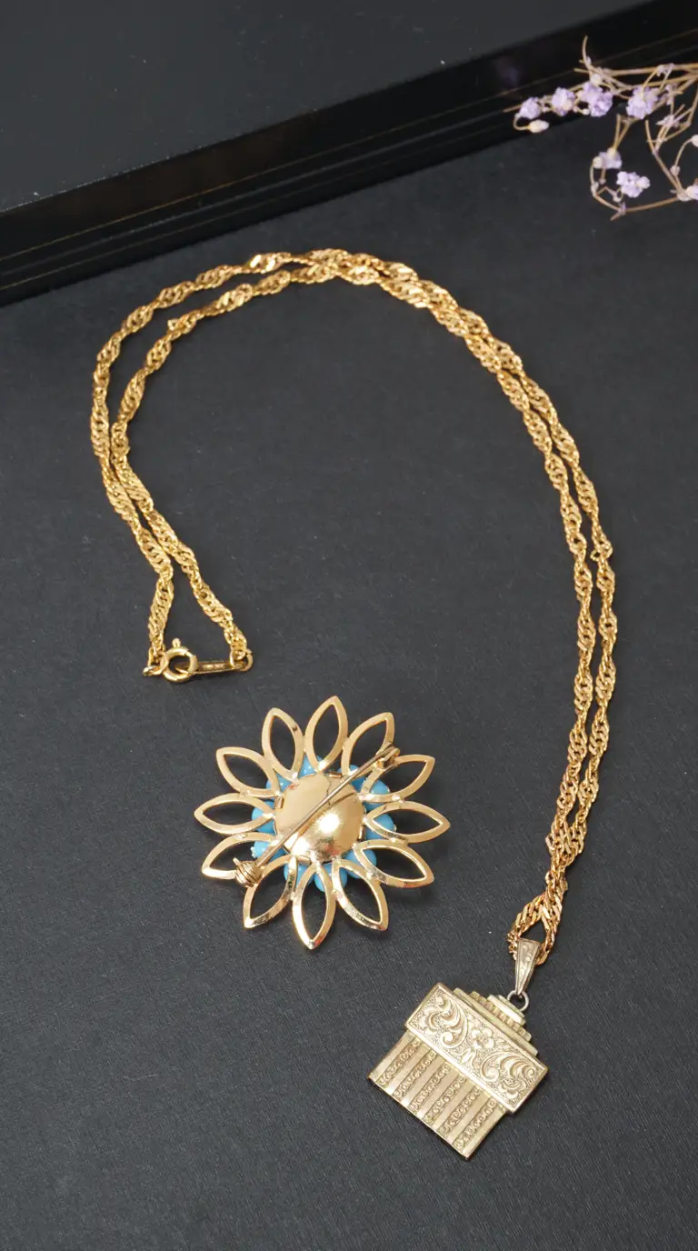 Vintage gold tone necklace with ornate engraved rectangular pendant and a flower-shaped brooch with blue accents, shown on a dark background.