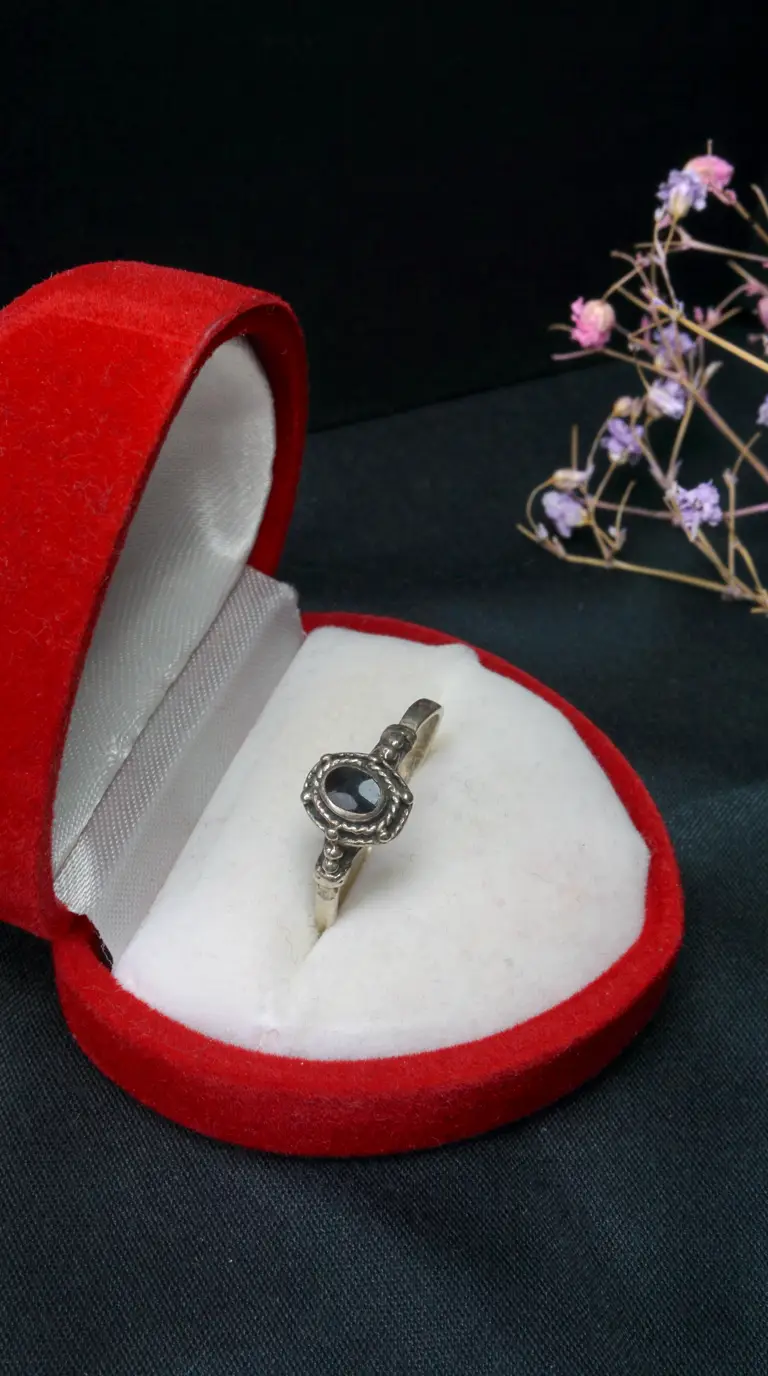 Vintage silver ring with ornate setting and black oval gemstone in an open red velvet jewelry box, with dried flowers.