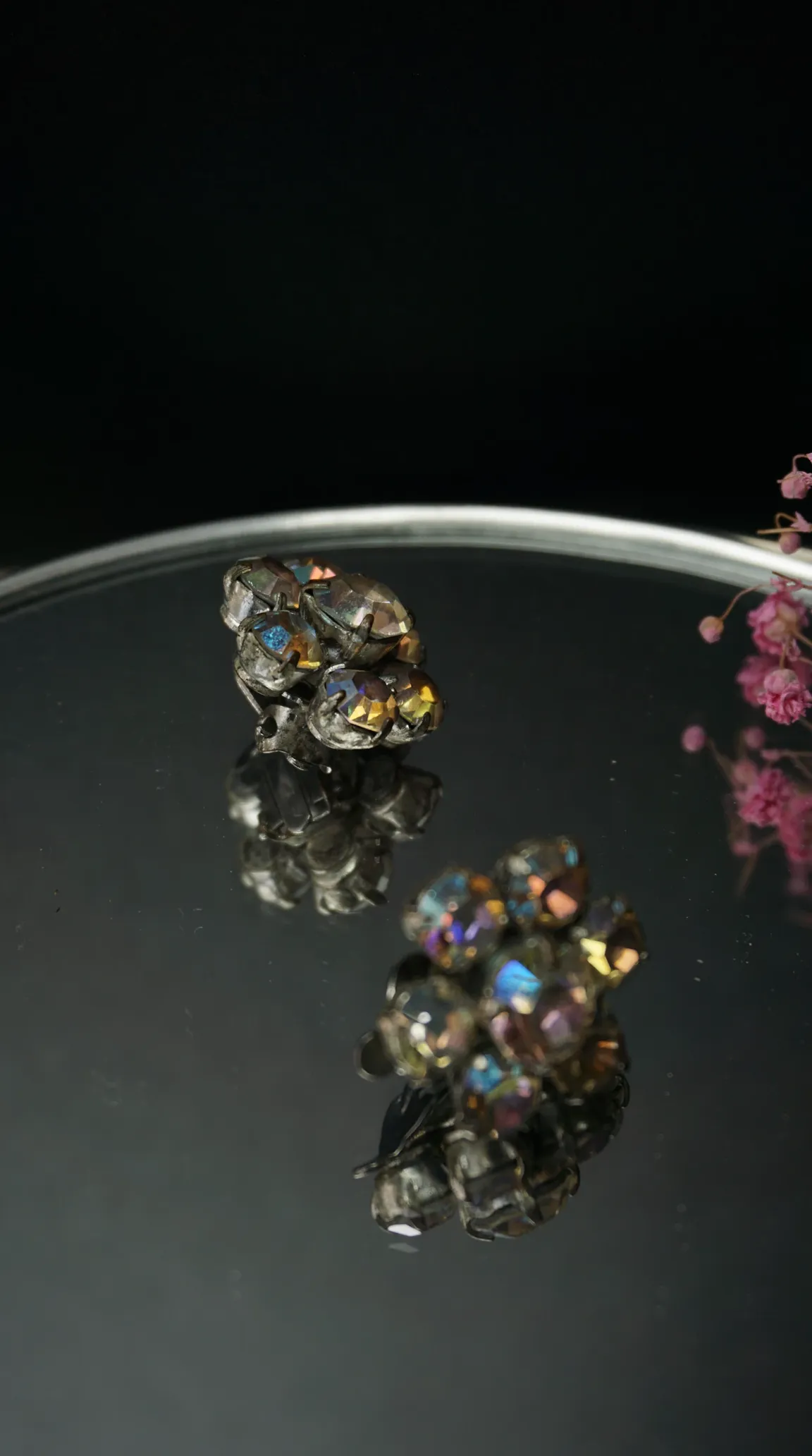 Vintage aurora borealis rhinestone cluster clip-on earrings, silver tone settings, on a mirrored surface with pink dried flowers.