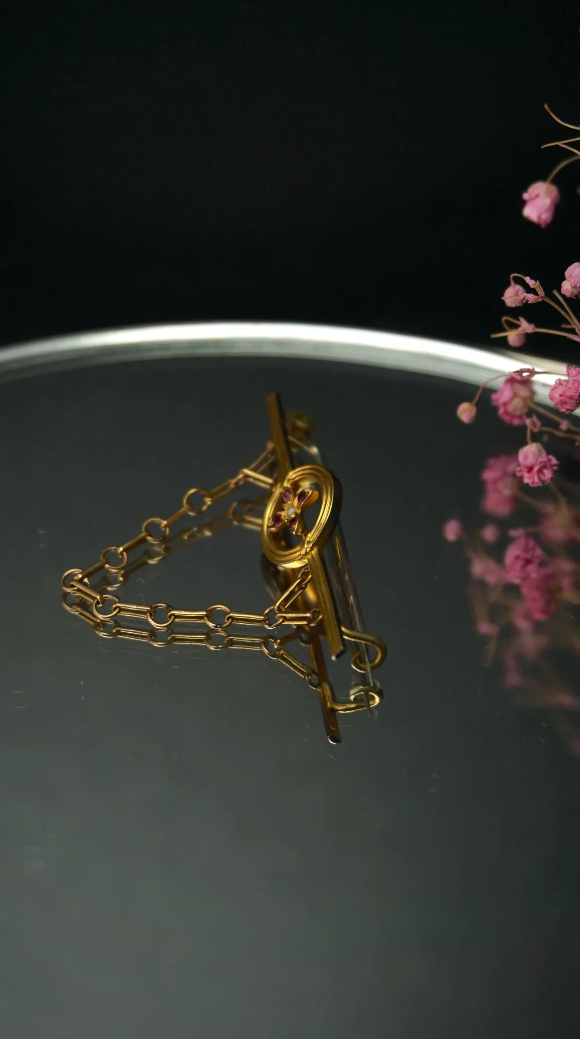 Vintage gold tie bar with chain, ruby and pearl flower design, on dark reflective surface.