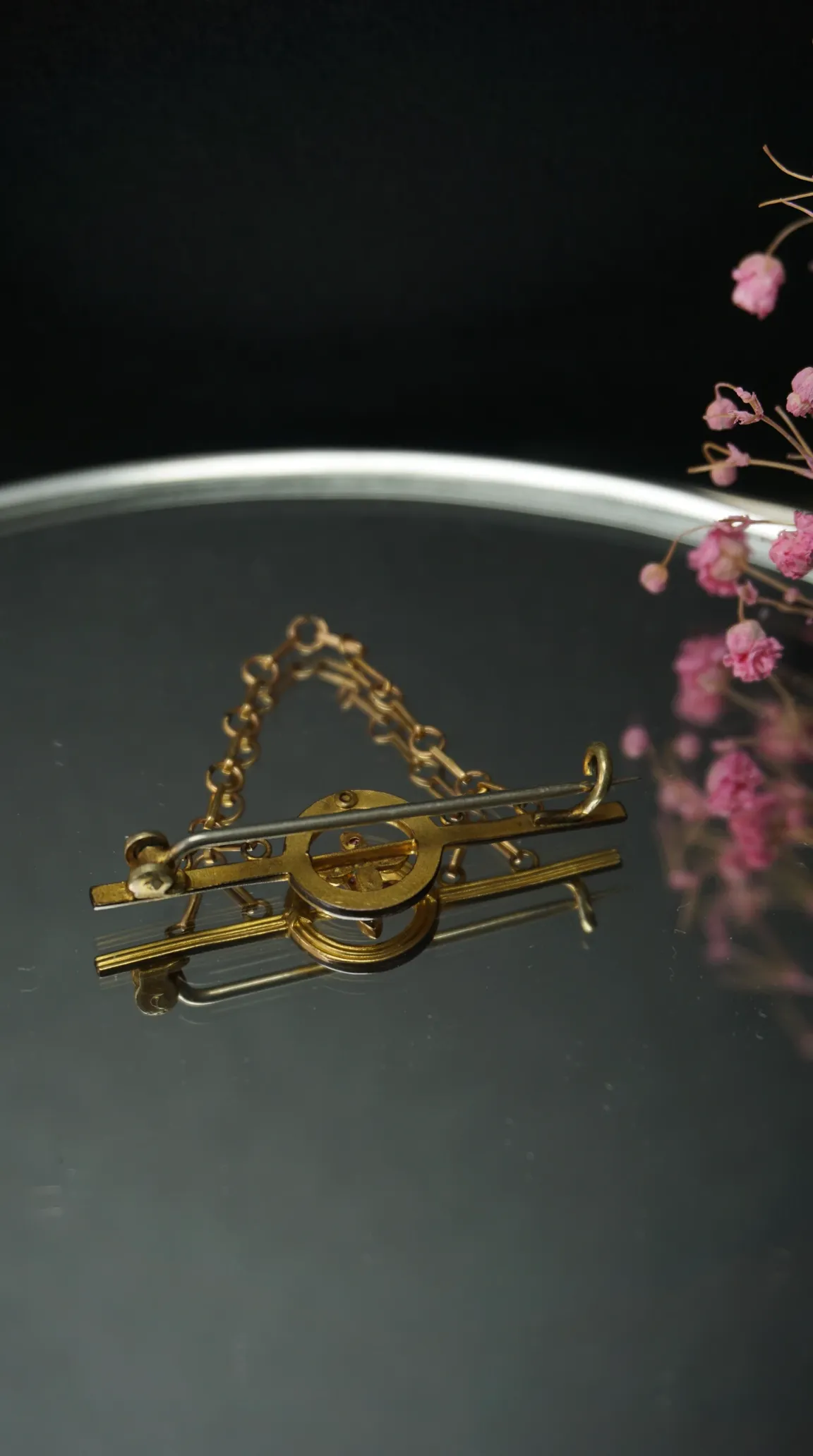 Vintage gold-tone ornate brooch with safety chain, displayed on a reflective surface with pink floral accents.