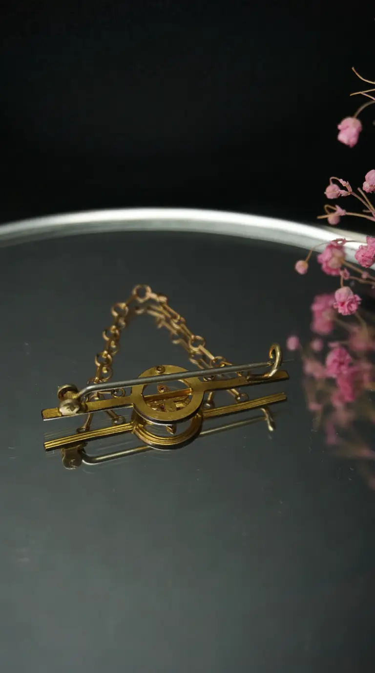 Vintage gold-tone ornate brooch with safety chain, displayed on a reflective surface with pink floral accents.