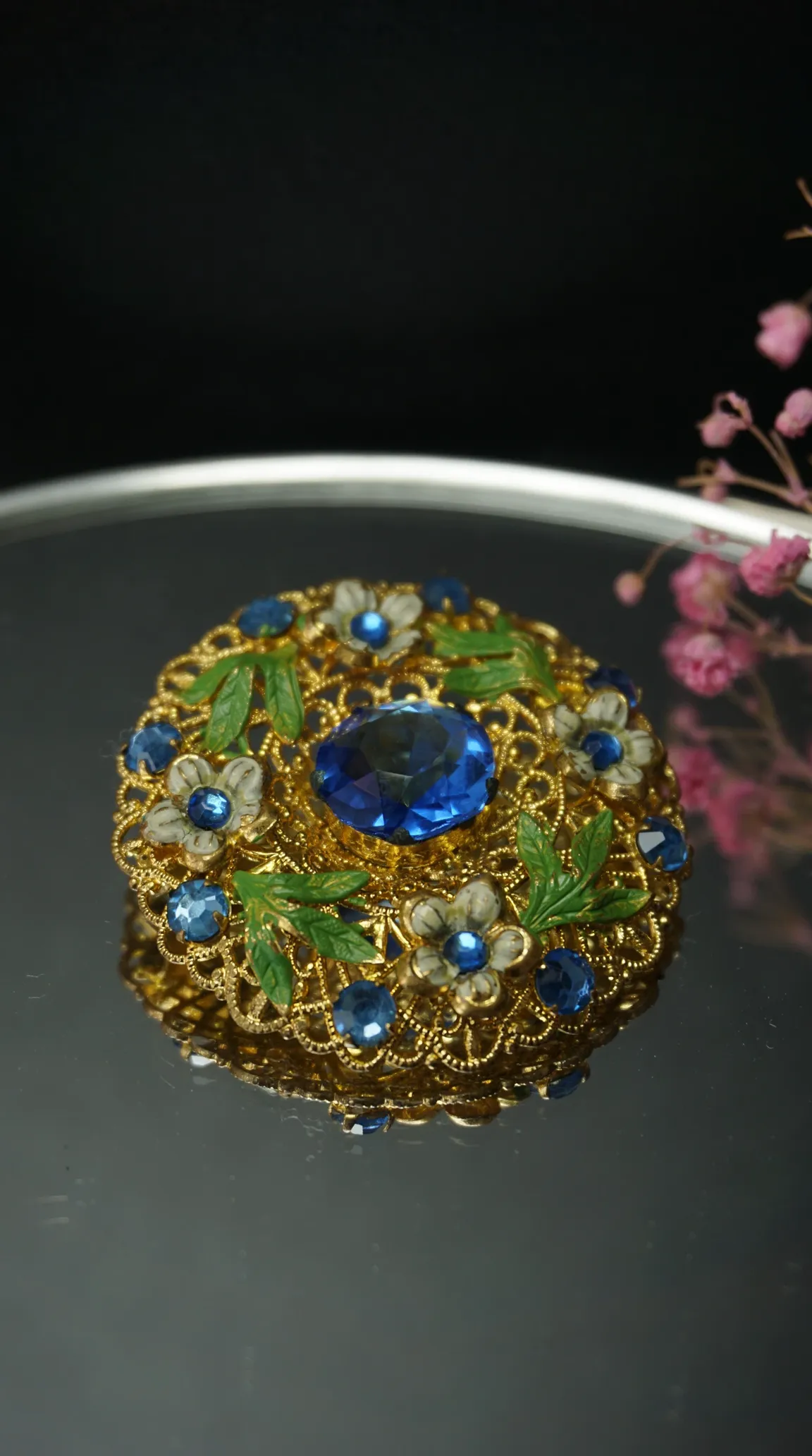 Vintage gold filigree brooch with large blue gemstone, smaller blue crystals, and white enamel flowers with green leaves.