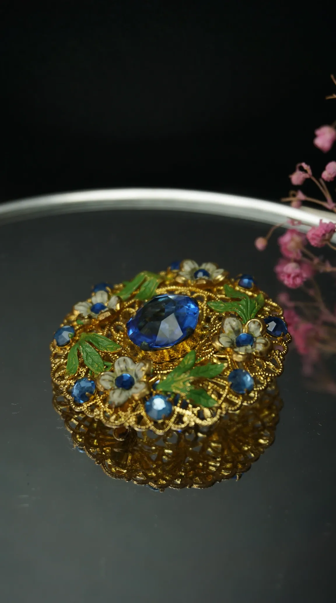 Vintage gold tone floral brooch pin with large blue gemstone, green enamel leaves, and white enamel flowers.