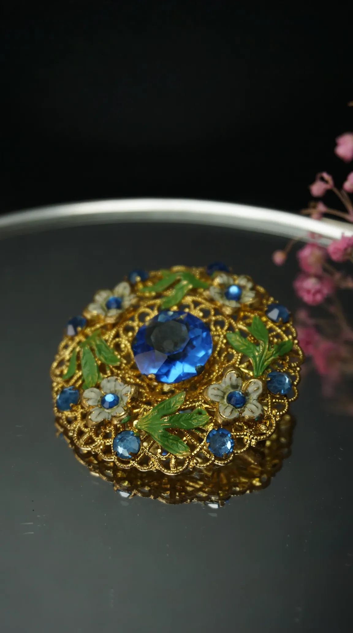 Vintage gold tone filigree brooch with large blue faceted crystal, smaller blue rhinestones, green enamel leaves, and white enamel flowers.