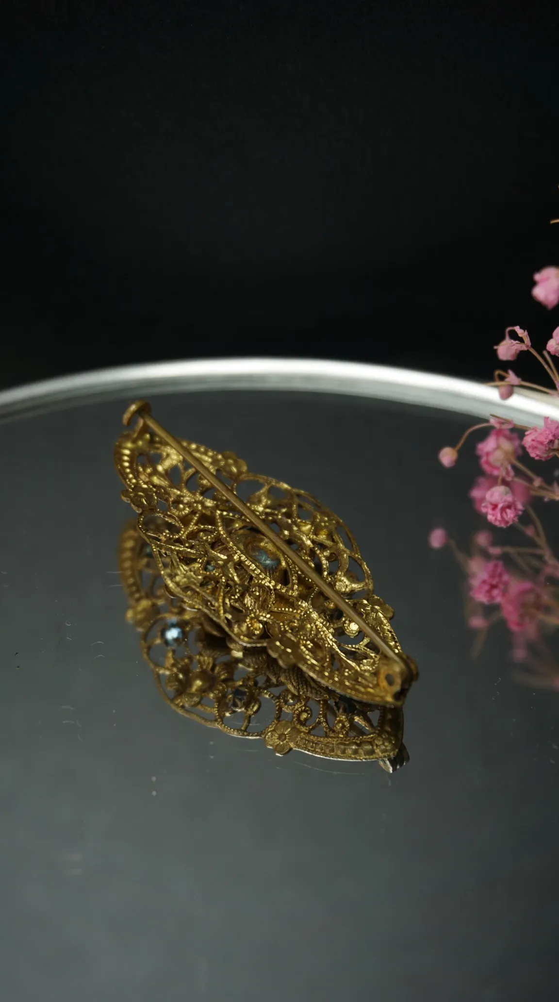Vintage gold-toned filigree brooch showing its reverse side with pin clasp on a reflective surface with pink flowers.