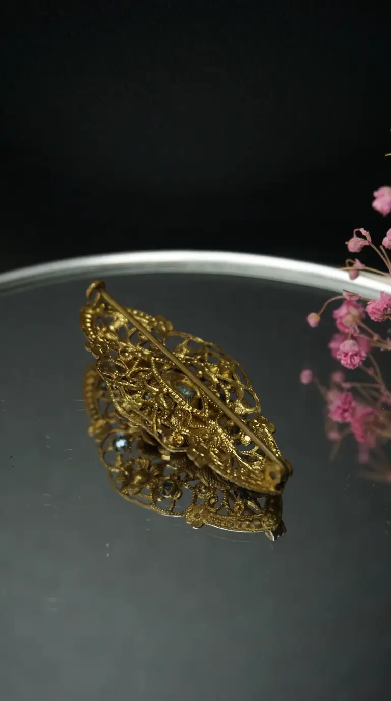Vintage gold-toned filigree brooch showing its reverse side with pin clasp on a reflective surface with pink flowers.