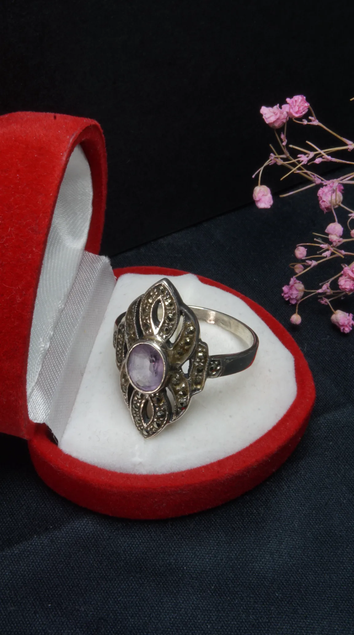 Vintage Art Deco style silver-toned ring featuring an oval lavender amethyst gemstone surrounded by shimmering marcasite accents, presented in an open red heart-shaped jewelry box.