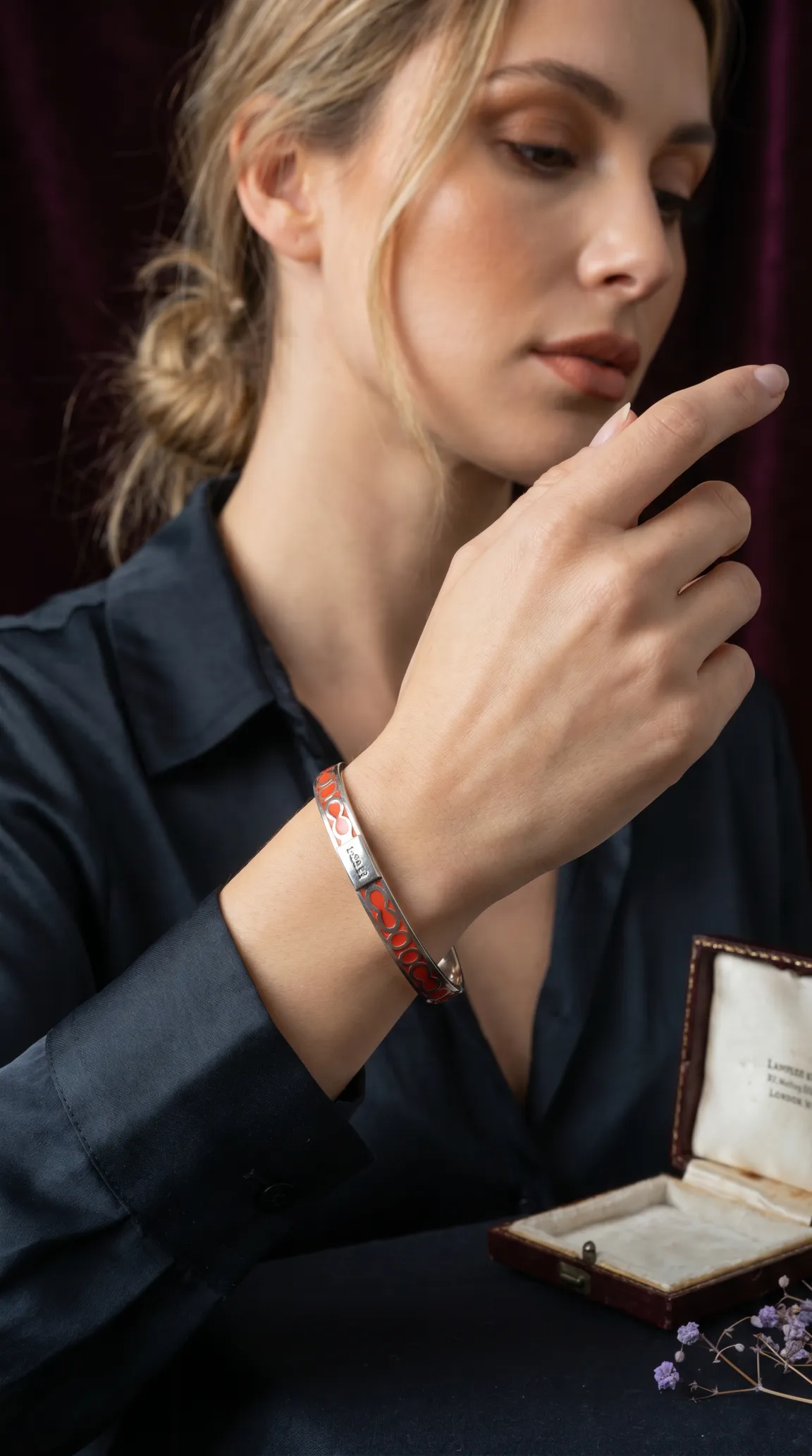 Vintage silver and red enamel bracelet with interlocking pattern and 'Jaeger' inscription on woman's wrist, next to an antique jewelry box.