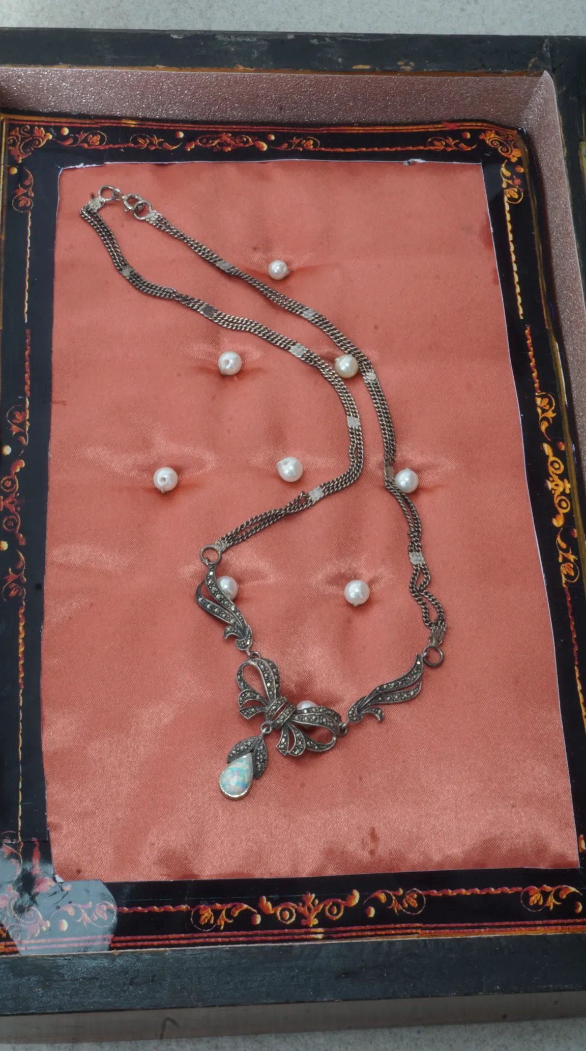 Vintage silver marcasite bow necklace with teardrop opal pendant, displayed on pink velvet with scattered pearls in an ornate antique box.
