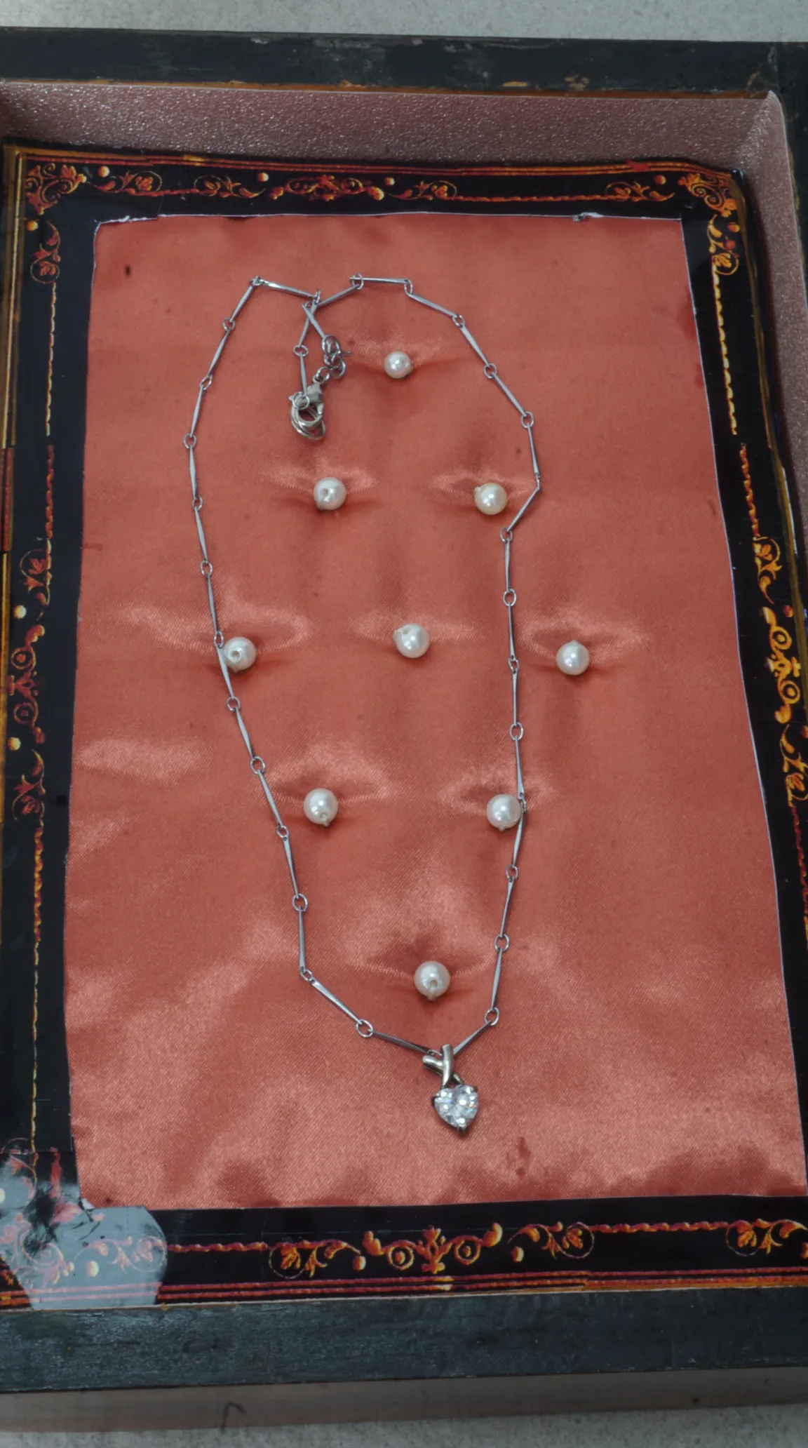 Vintage silver chain necklace with clear crystal heart pendant, displayed on coral satin with loose pearls in an ornate black and gold box.
