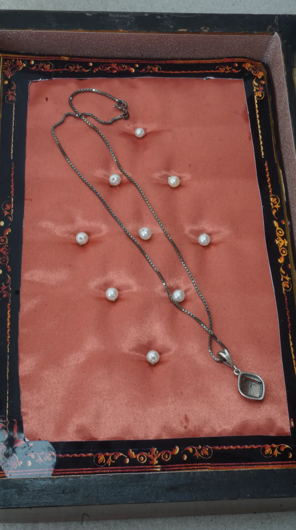 Vintage oxidized silver box chain necklace with geometric kite pendant, displayed on a peach satin jewelry cushion with small pearl details inside an ornate black and gold box.