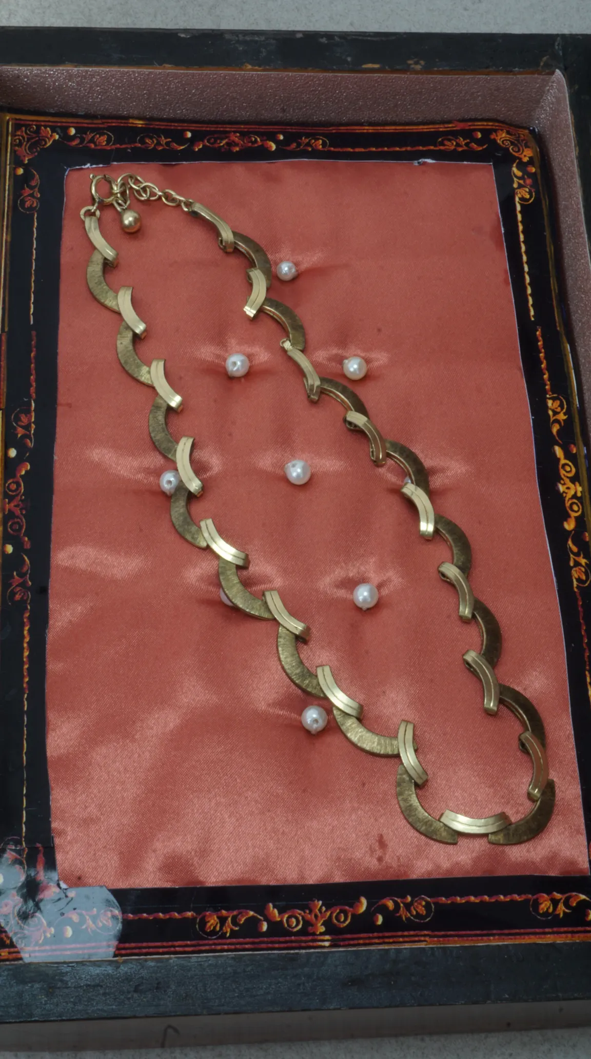 Vintage gold tone crescent link necklace with brushed finish, displayed on peach satin fabric with scattered pearls, inside a decorative black and gold jewelry box.