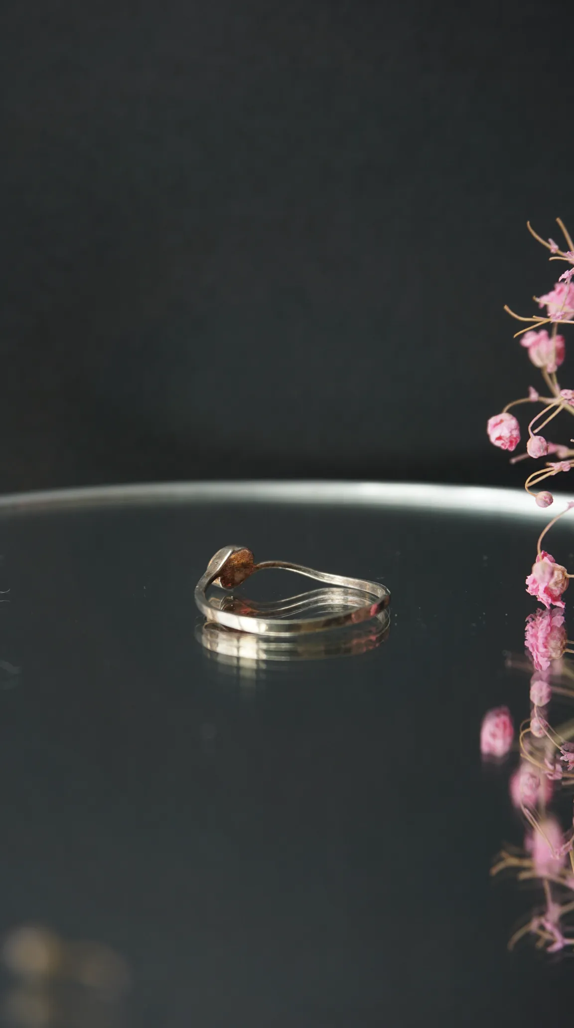Vintage sterling silver ring with reddish-brown gemstone on a dark reflective surface with dried pink flowers.