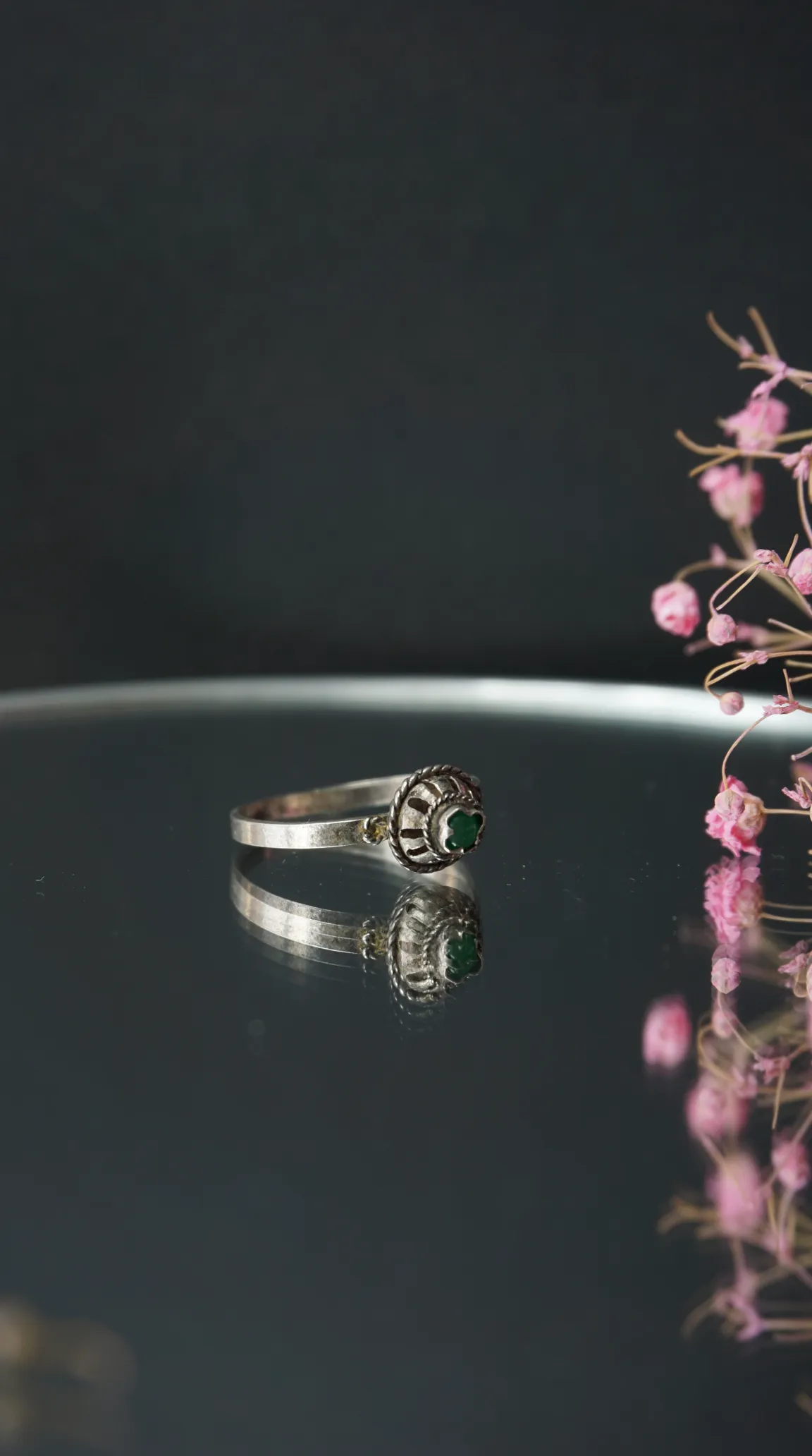 Vintage silver ring with ornate setting and emerald green gemstone reflecting on a dark surface, with pink flowers.