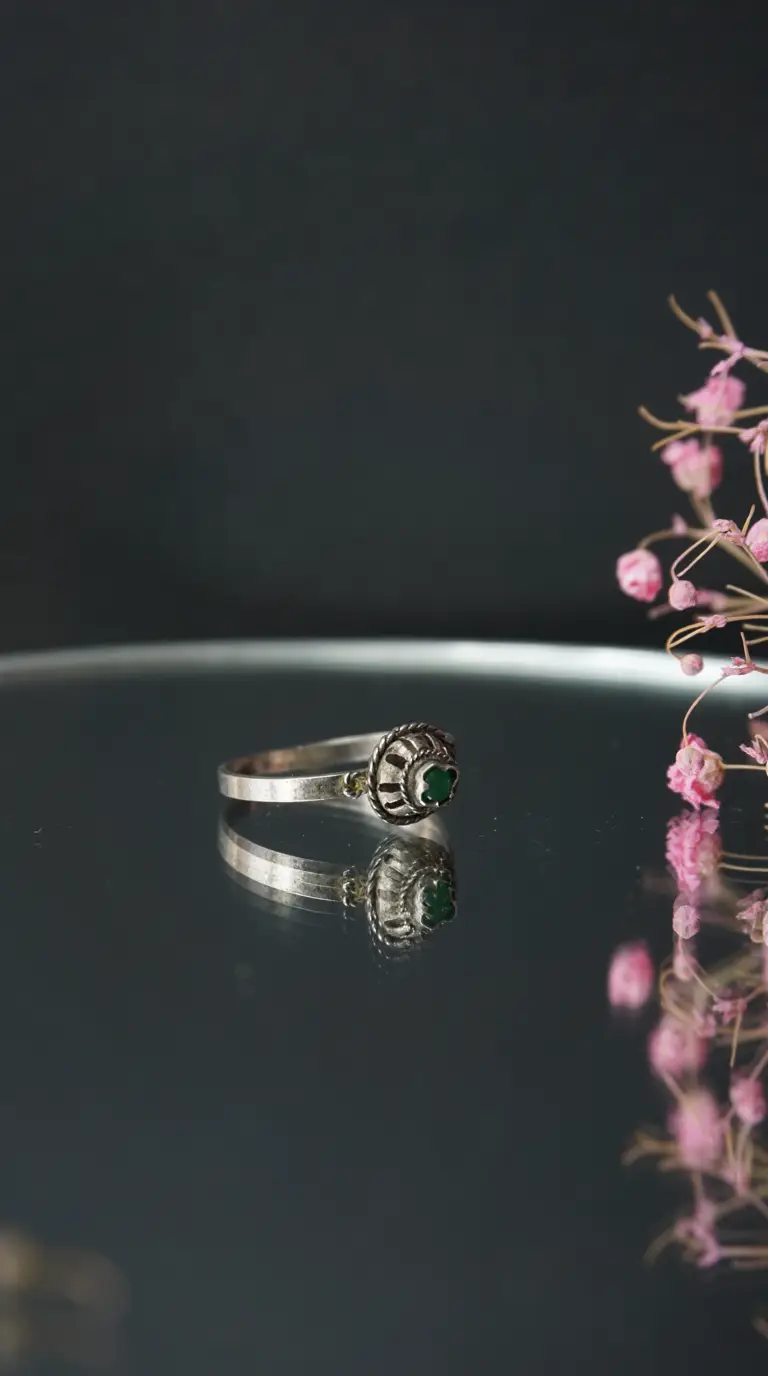 Vintage silver ring with ornate setting and emerald green gemstone reflecting on a dark surface, with pink flowers.