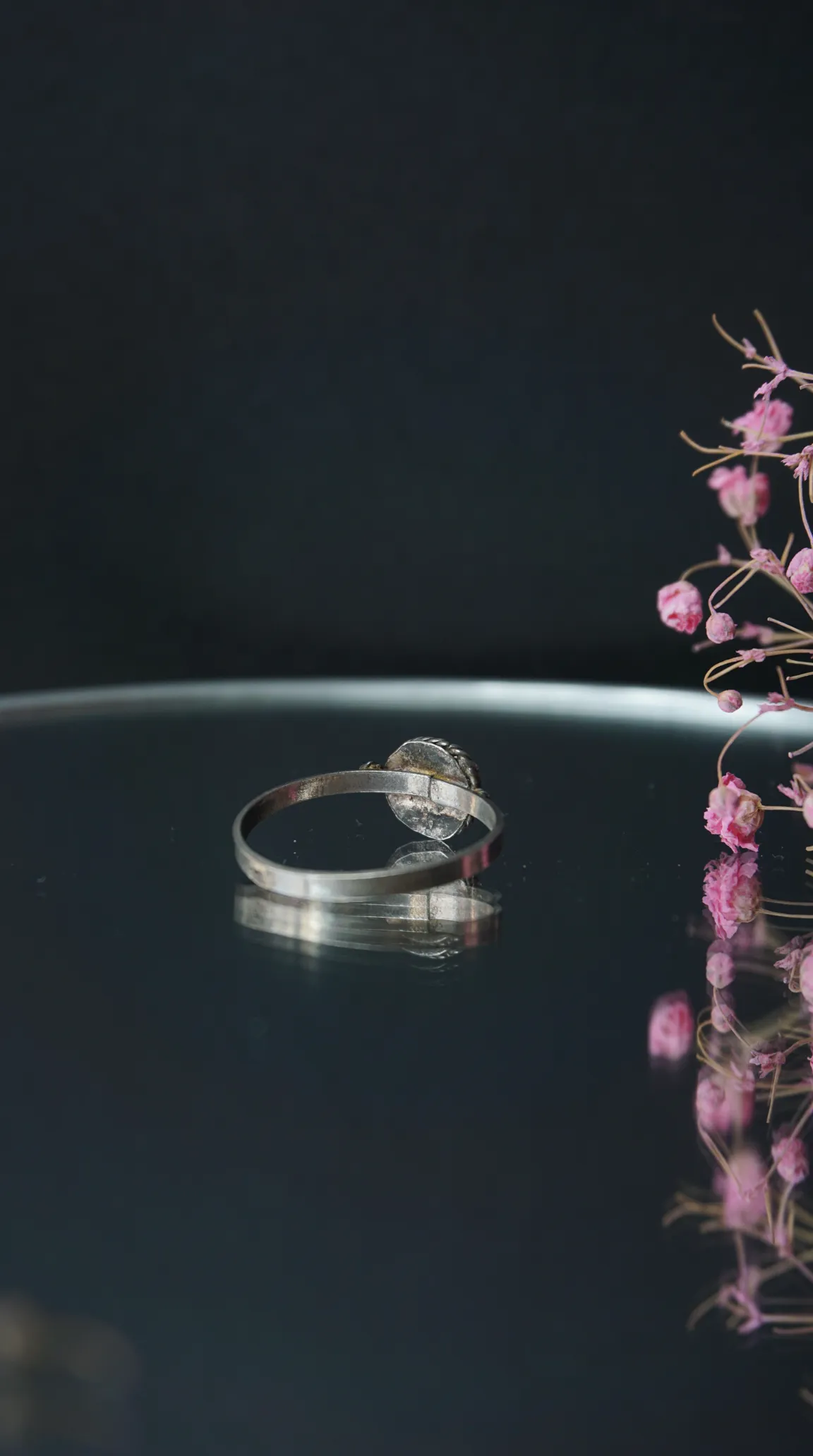 Vintage silver-toned ring with detailed setting on a dark reflective surface, accented by dried pink flowers.