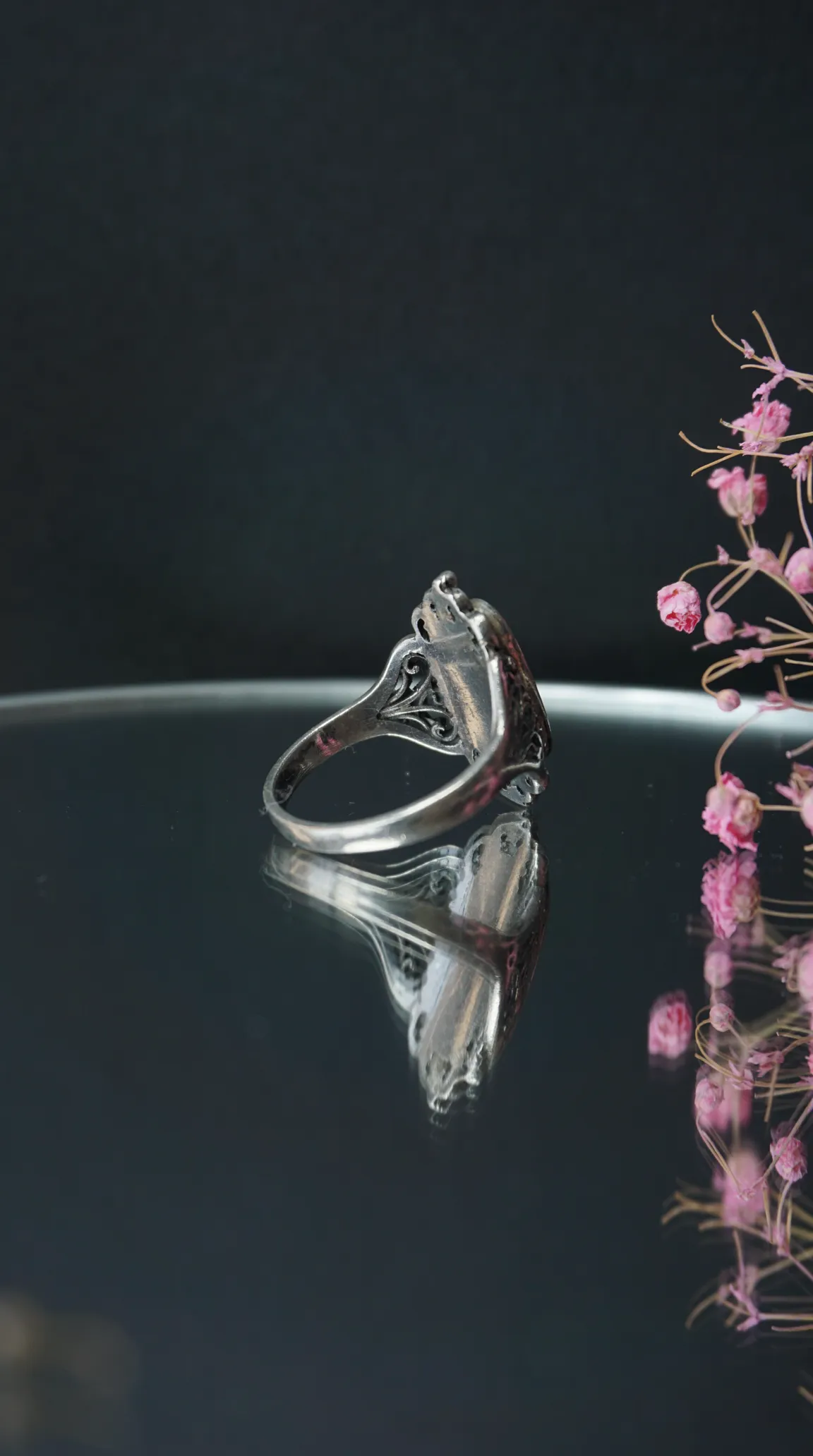 Vintage silver filigree ring with ornate details reflected on a dark surface with pink dried flowers.