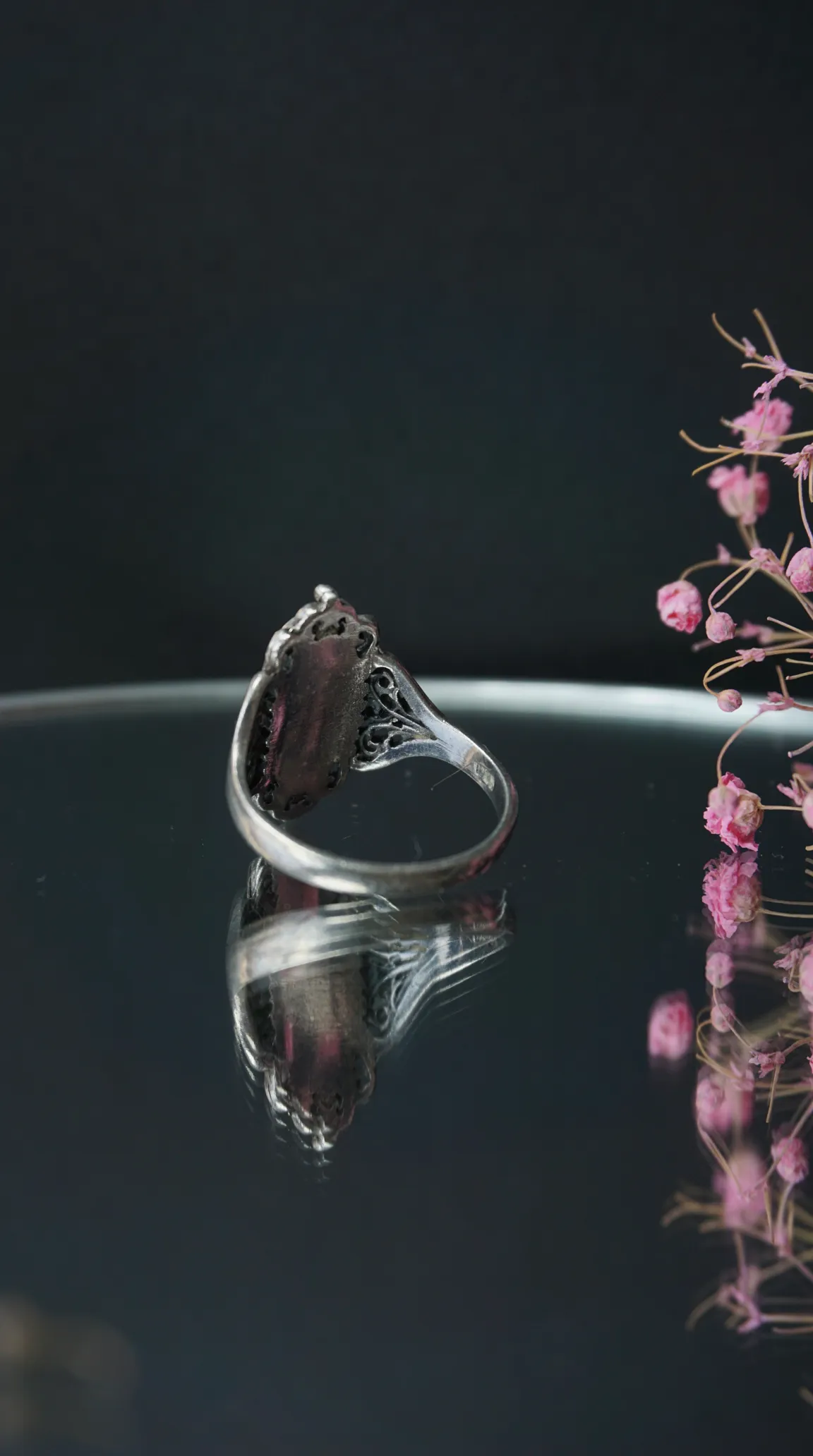 Vintage silver filigree ring with dark oval gemstone, reflected on glossy surface, pink dried flowers.