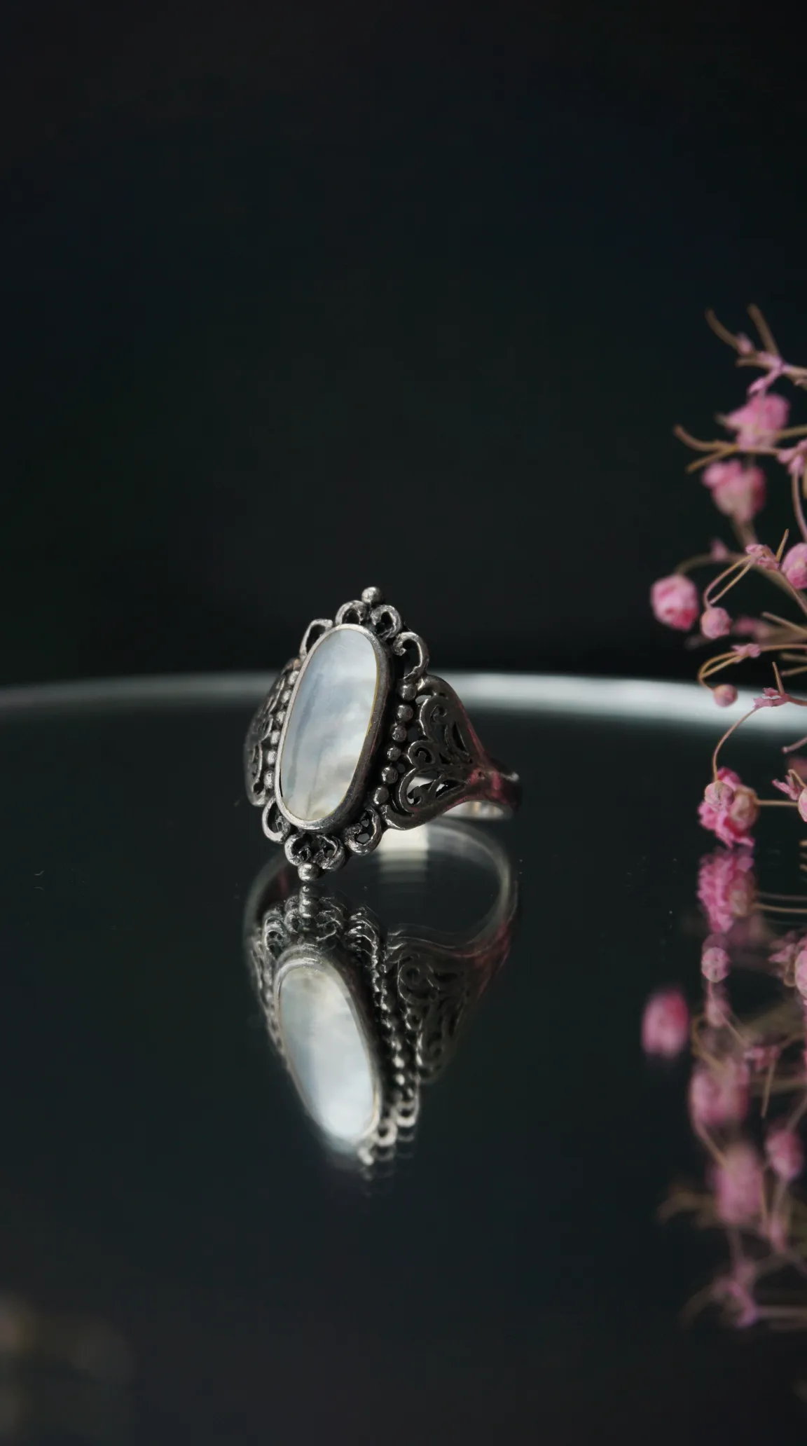 Vintage sterling silver ring with oval mother-of-pearl gemstone and ornate filigree detail on a reflective dark surface.