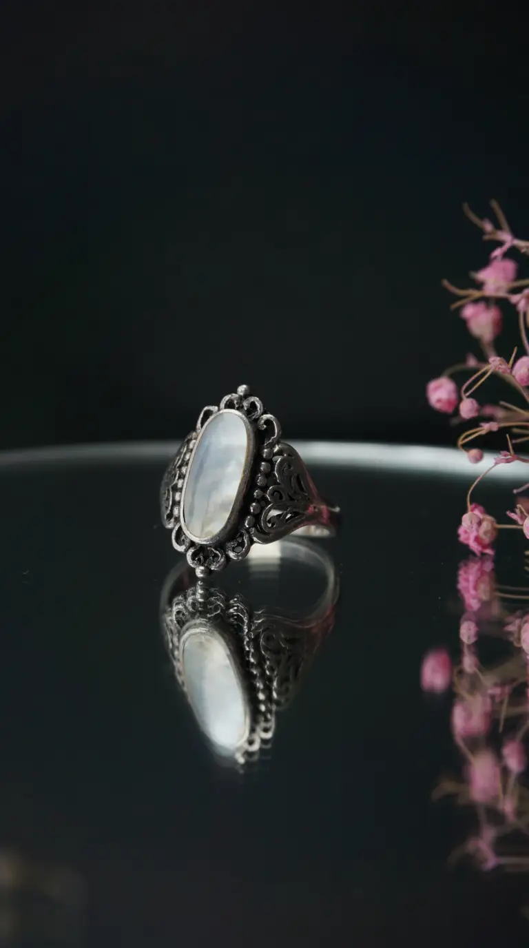 Vintage sterling silver ring with oval mother-of-pearl gemstone and ornate filigree detail on a reflective dark surface.