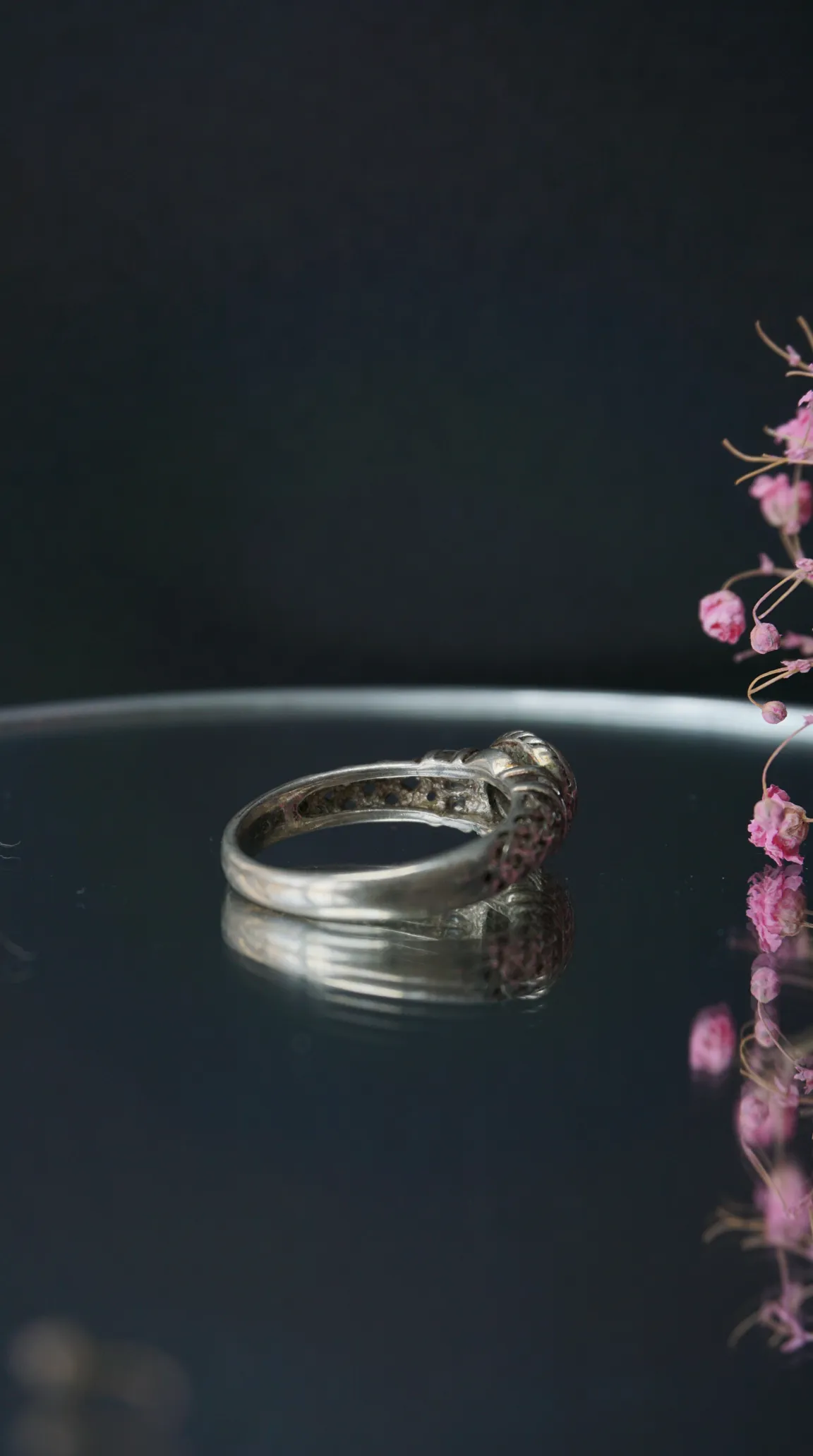 Vintage silver ring with intricate band design and stone setting, reflecting on a dark surface, with pink flowers.