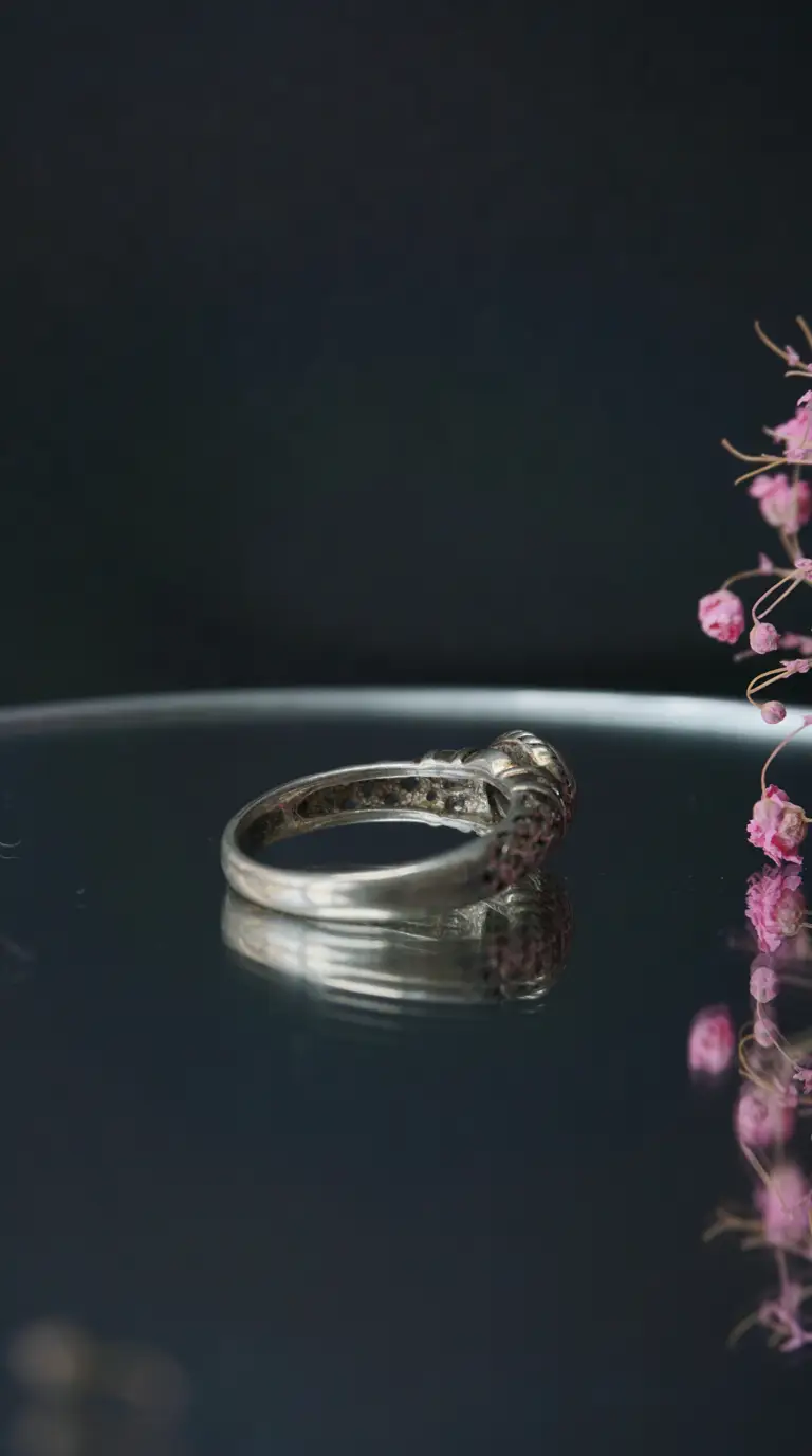 Vintage silver ring with intricate band design and stone setting, reflecting on a dark surface, with pink flowers.
