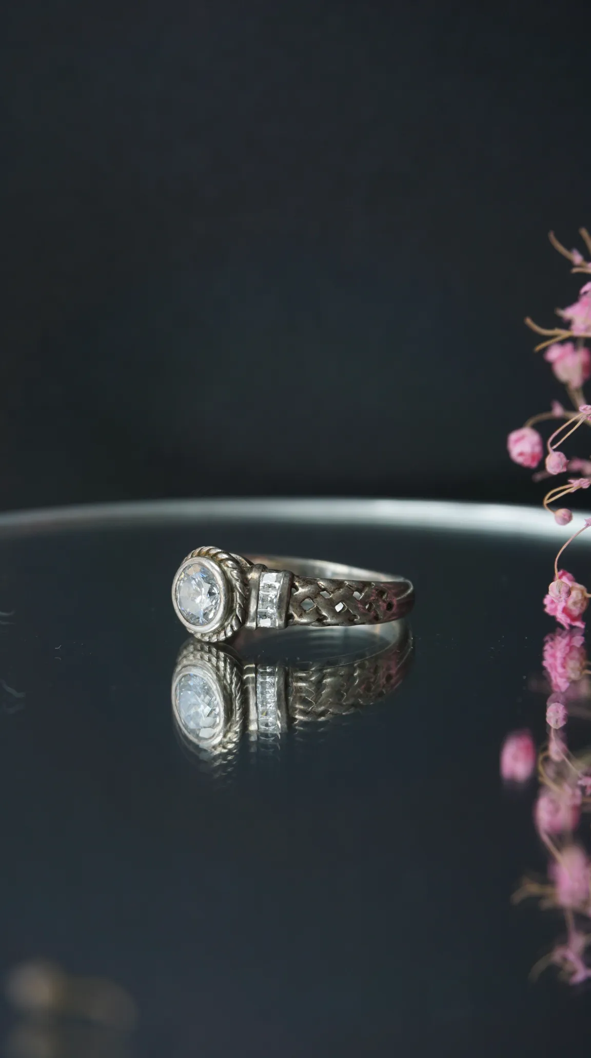 Vintage silver ring with a large round clear stone, two rectangular side stones, and an intricate interwoven band, reflected on a dark surface.