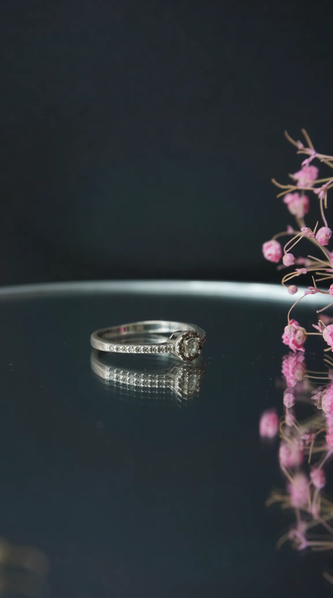 Vintage silver diamond halo ring with pavé band, reflected on a dark surface with delicate pink flowers.